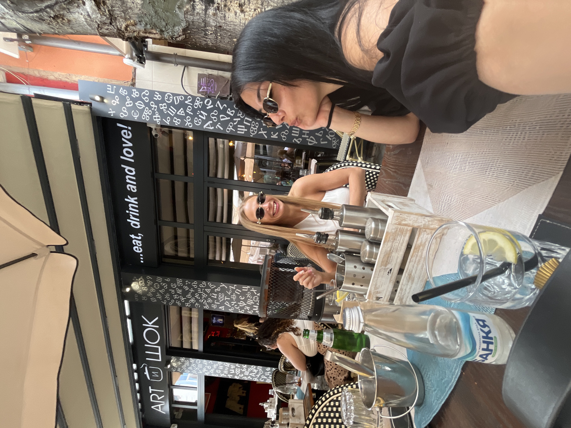 The image shows two women at an outdoor table outside of the ART WOK restaurant in Plovdiv, Bulgaria. One woman with dark hair is facing the camera with her head resting on her hand. She is wearing a black top and sunglasses. The other woman with blonde hair is also facing the camera with sunglasses. She is wearing a white tank top. The table has a textured placemat and contains condiments, glasses, and a bottle of water labeled "BAHKA" and a green bottle, likely mineral water. The restaurant has large glass windows and a sign that reads "...eat, drink and love!" above "ART WOK" in Cyrillic letters. The storefront is decorated with white and black abstract shapes. A canopy provides shade. Other patrons are visible inside the restaurant.