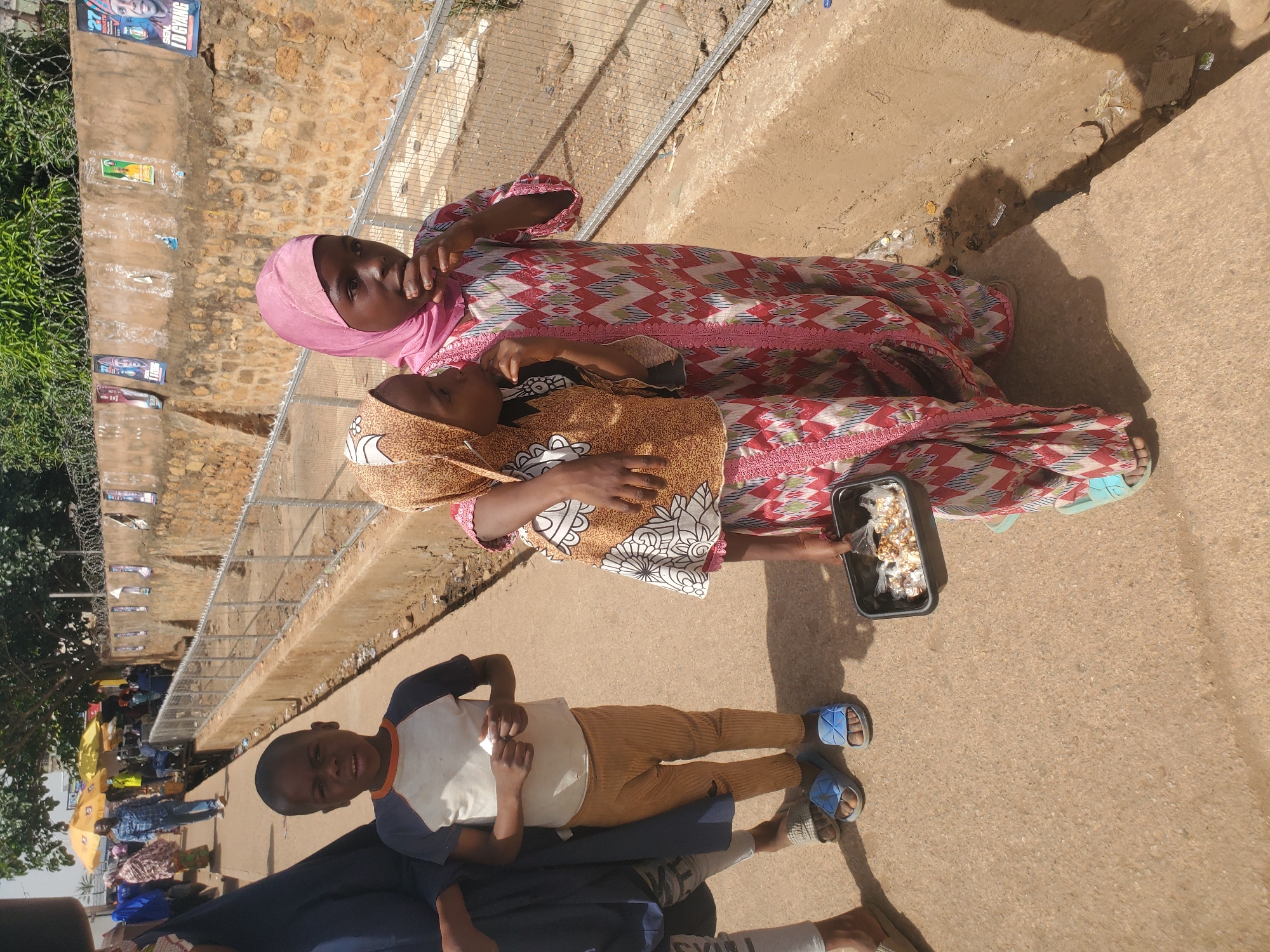 This image, taken in Bukuru, Nigeria, captures an outdoor scene on a bright, sunny day. In the foreground, two young girls or women are visible. One, positioned on the right, wears a pink hijab and a long, flowing dress with intricate red, white, green, and blue geometric patterns. She holds a black rectangular container filled with popcorn or a similar snack. To her left, a younger girl, likely a child, wears a brown and white patterned top with a head covering, looking towards the camera.
Further to the left, a young boy stands, wearing a white and dark blue t-shirt, light brown trousers, and blue slide sandals, offering a faint smile to the camera. In the background on the left, other individuals are seen, some under yellow umbrellas, suggesting a street market or a busy public space.
The setting includes a dirt and paved pathway. A metal fence topped with barbed wire runs along the left side, separating the pathway from a lower area with green vegetation. Behind this fence, a stone wall is visible, on which several posters are affixed. One distinct poster at the top left prominently displays the text "SEYI" and "TI CHANG," likely a political advertisement. The strong sunlight casts visible shadows, indicating it is daytime. The scene portrays a lively community environment.