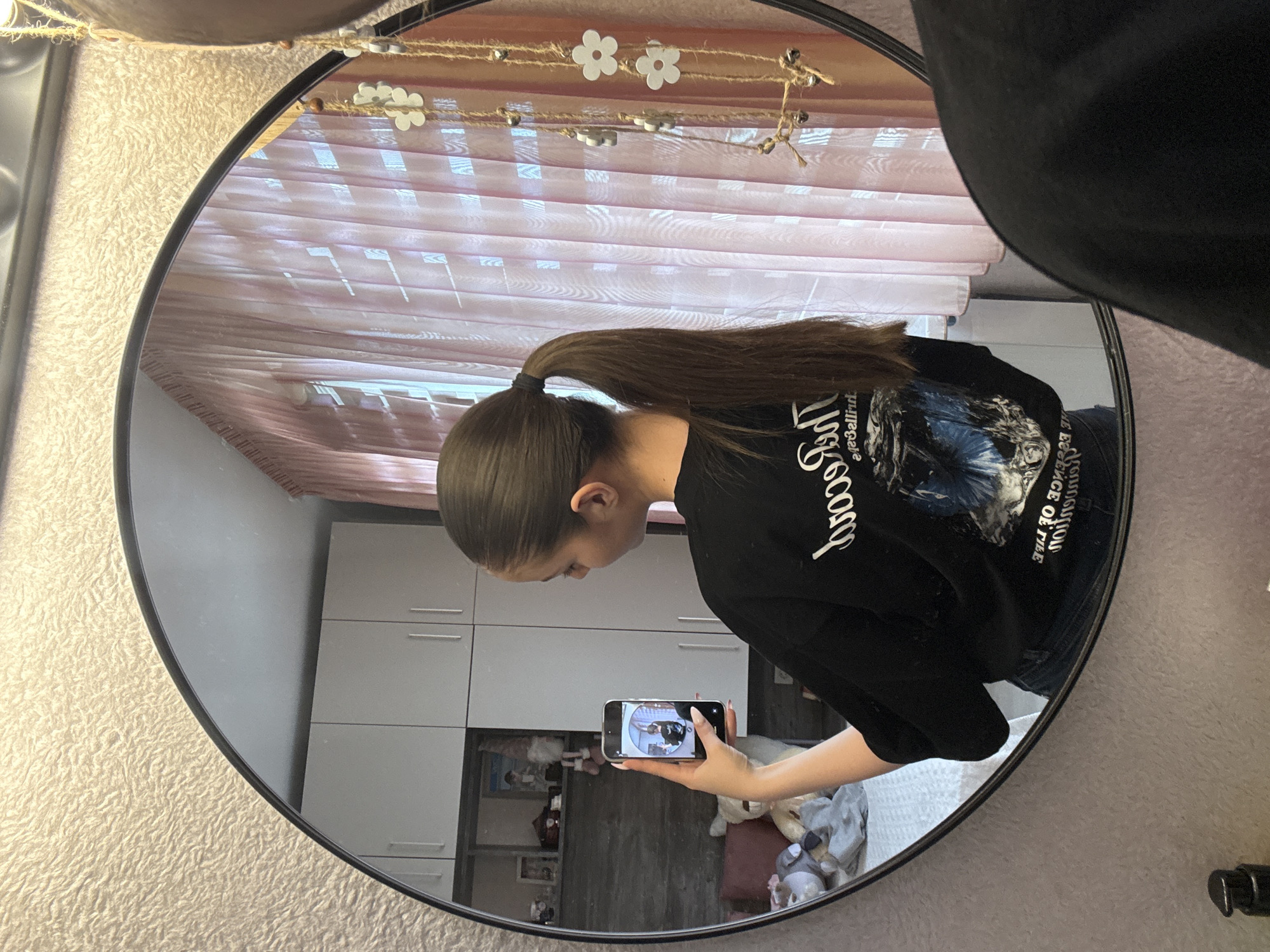 A young woman is reflected in a round mirror. She holds a smartphone in her hand, appearing to be taking a photograph of herself in the mirror. The woman has dark hair pulled back in a ponytail and is wearing a black sweatshirt with white lettering. The background shows a room with a wardrobe and a window with sheer, light pink curtains. The mirror has a string of decorative flowers above. The setting appears to be in a residential space in Bulgarovo, Bulgaria, based on the available information.