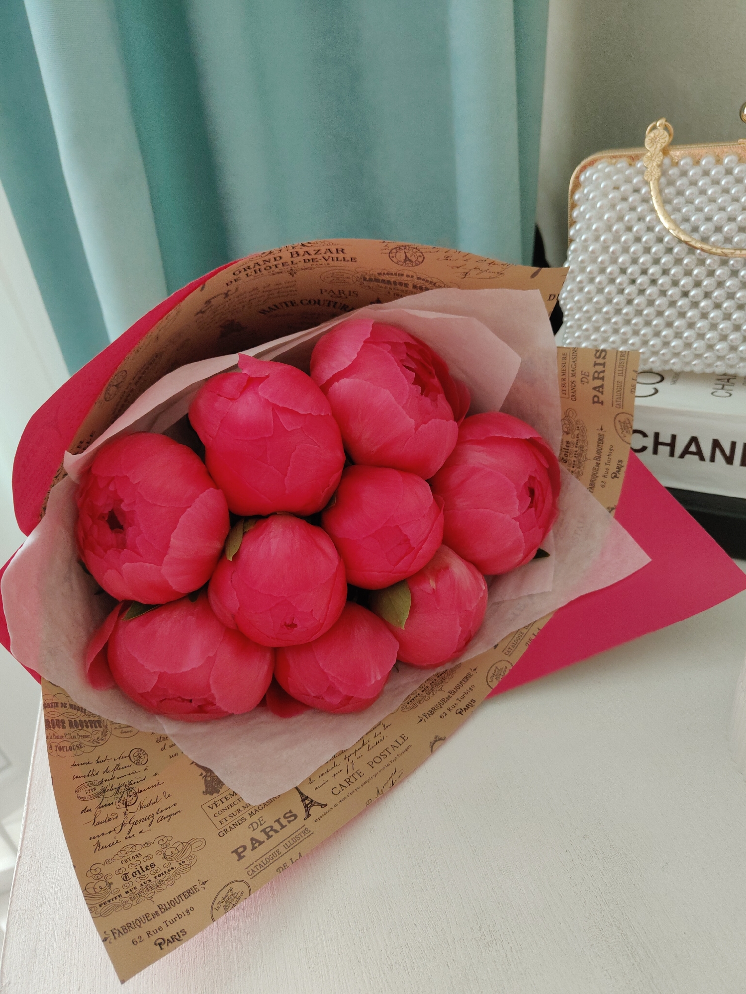A vibrant bouquet of tightly budded pink peonies is prominently displayed, wrapped in a combination of magenta paper and beige kraft paper with a vintage French theme. The flowers are arranged closely together, some beginning to unfurl their petals. Thin white tissue paper is also part of the wrapping, providing a soft contrast.
The bouquet rests on a white, textured surface, possibly a table or shelf. In the background, to the right, a white pearl-beaded handbag with a gold handle adds a touch of elegance. Behind the handbag, a stack of books is partially visible, with the word "CHANEL" clearly readable on the spine of one book. To the left and top of the image, light teal or greenish-blue curtains with vertical stripes provide a soft, draped backdrop, suggesting an indoor setting bathed in natural light.
The beige wrapping paper is covered with various French-themed text and small illustrations, reminiscent of old Parisian postcards or advertisements. Visible text includes "GRAND BAZAR DE L'HOTEL-DE-VILLE", "HAUTE COUTURE", "PARIS" (repeated multiple times), "CARTE POSTALE", "FABRIQUE DE BIJOUTERIE", "62 Rue Turbigo", "CATALOGUE ILLUSTRE", "GRANDS MAGASINS", and "VÊTEM CONFECTI ET SUR MI". Small drawings, including what appears to be the Eiffel Tower, are interspersed among the text. The overall aesthetic suggests a delicate and refined scene, possibly a gift or a decorative arrangement in a room.
