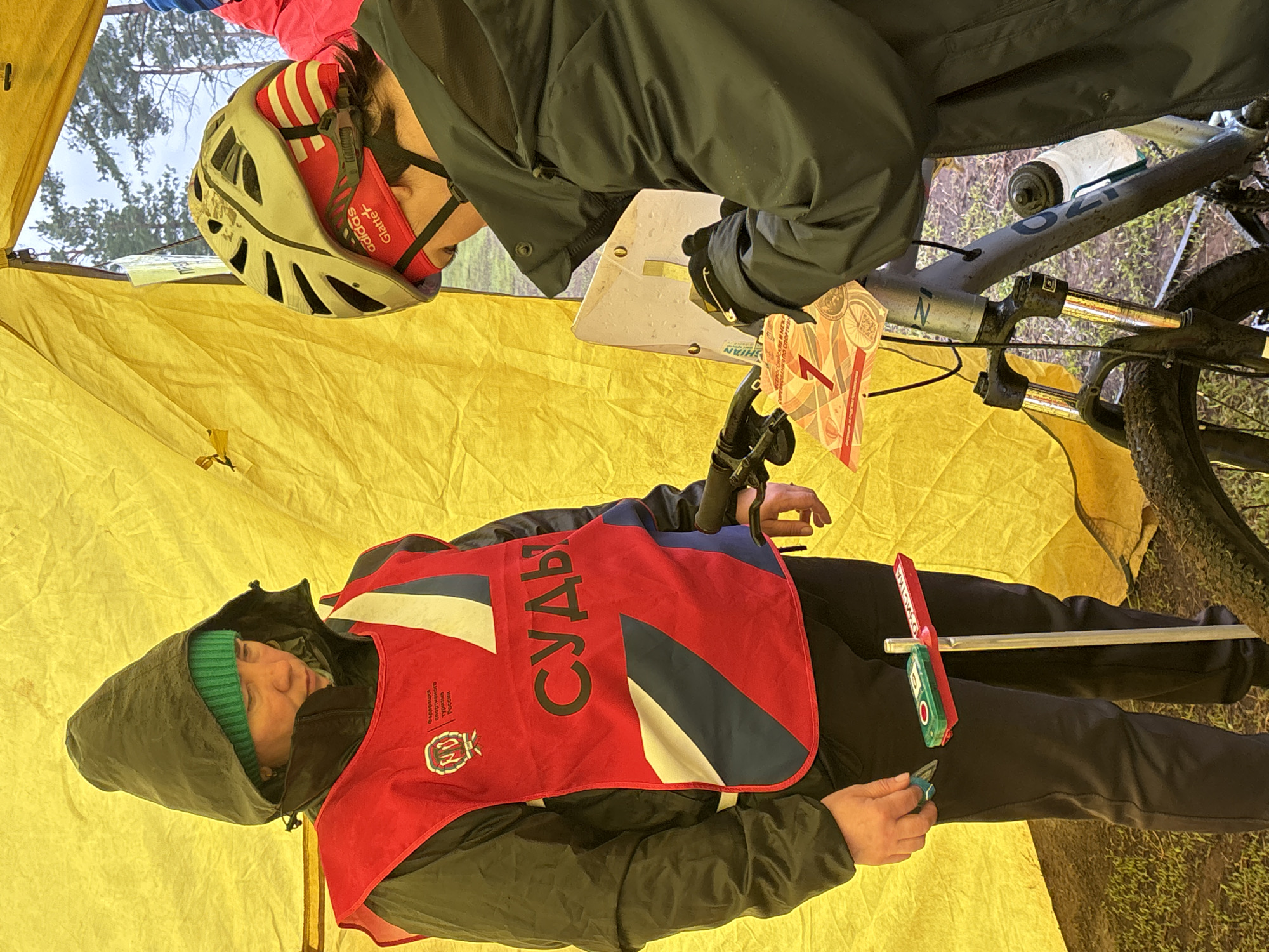 The image, likely captured in Orenburg, Russia, depicts a yellow tent interior. Inside, two individuals are present. One person, wearing a red vest with the word "СУДЬЯ" (Judge) in Cyrillic script and a green beanie, lies horizontally on the tent floor. The other person, wearing a helmet and dark jacket, stands beside a bicycle, holding a clipboard and writing. A number '1' marker is attached to the bicycle's handlebars.