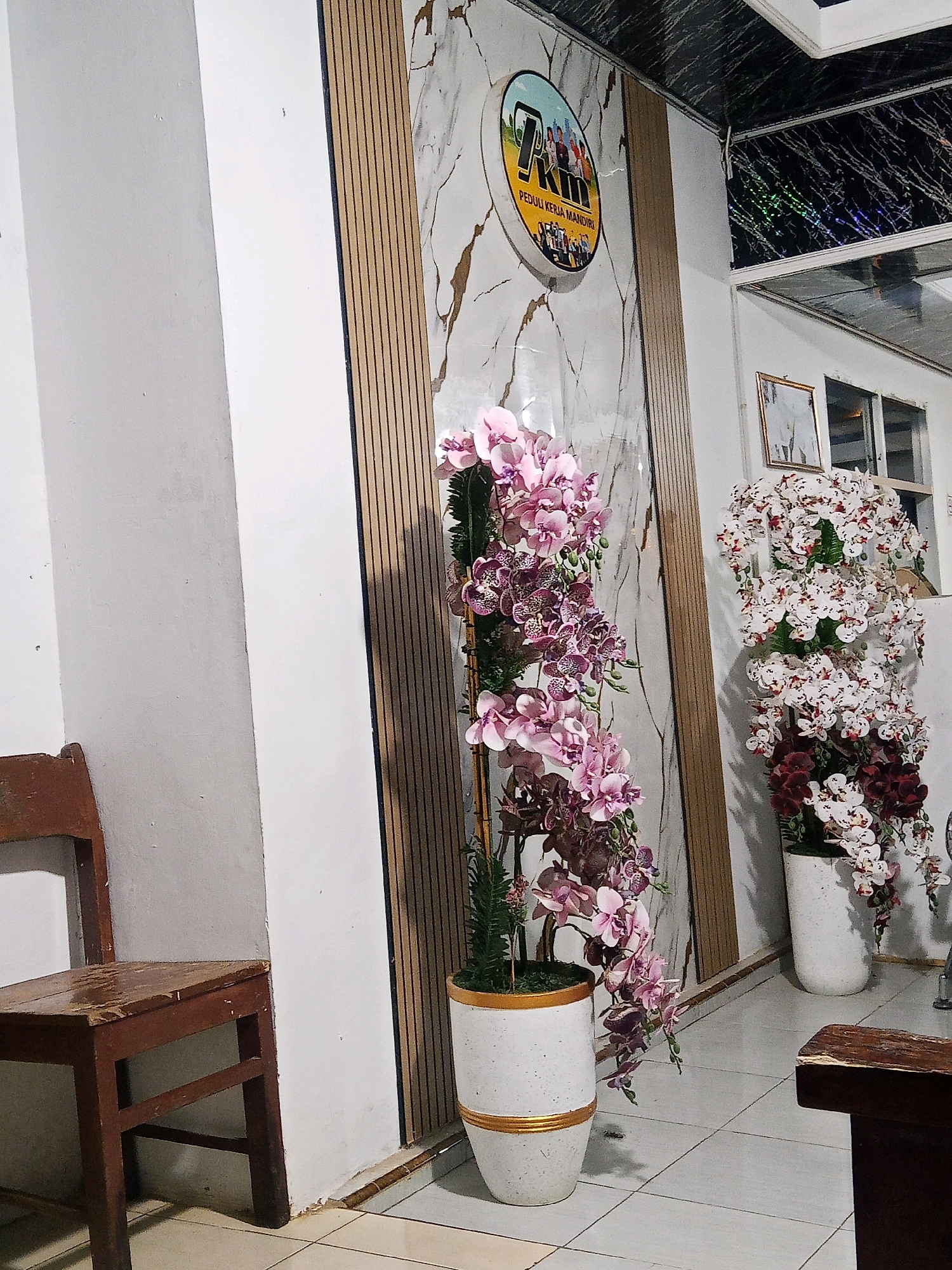 The image depicts an indoor scene with a focus on decorative floral arrangements and architectural elements. In the foreground, a weathered wooden chair sits on a tiled floor. To the right, a tall white pot contains a cascading display of pink and purple artificial orchids. Further to the right, another white pot holds white and deep red artificial orchids. Behind the orchids, a wall features a large marble-patterned panel with vertical wooden slats. A circular sign with a logo featuring people and text is mounted on this wall. The ceiling above has a dark, reflective surface with a marbled pattern and some holographic elements. The overall impression is of an indoor space, possibly a waiting area or a decorated corner of a building, likely in Lubuklinggau, Indonesia, suggested by the text on the sign.