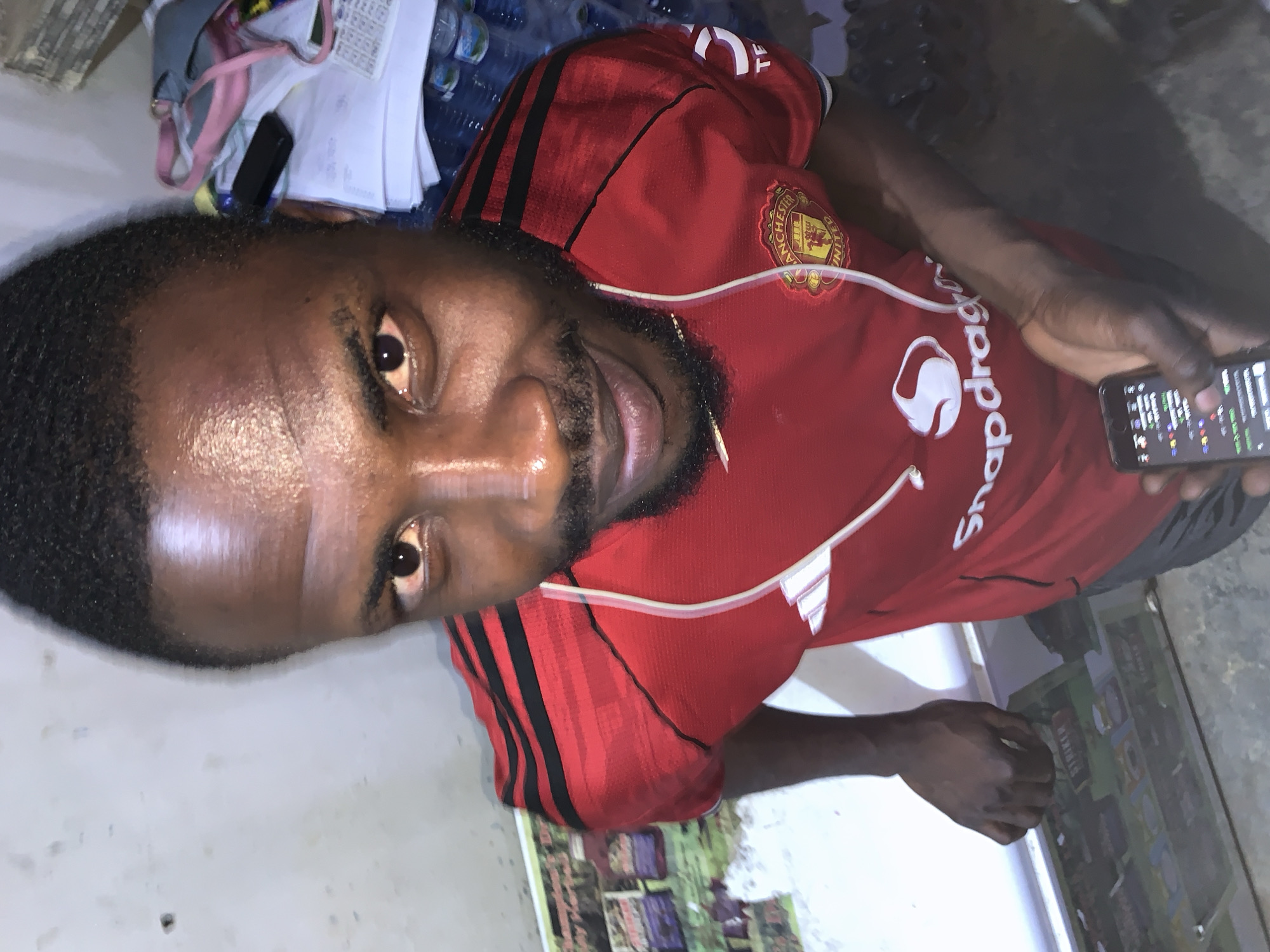 A man wearing a red Manchester United soccer jersey is looking at the camera while holding a smartphone. He is in a room with a cluttered desk. He is located in New Stadium, Nigeria.