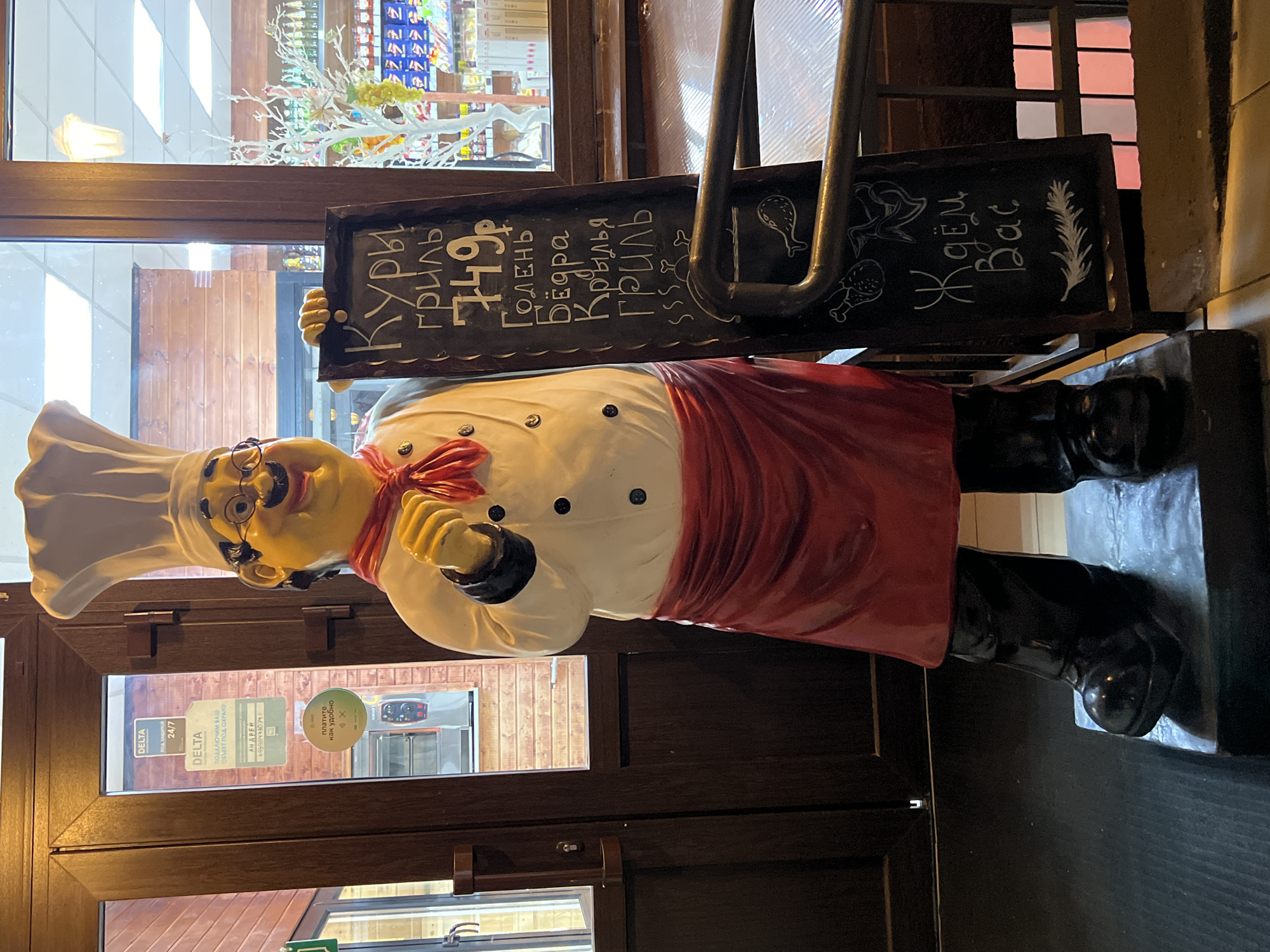 A full-body, life-sized statue of a chef wearing a white chef's hat, red scarf, white uniform with black buttons, and a red apron stands in front of a wooden door and a window. The chef is holding a chalkboard sign with a menu written in Russian. The menu lists "Кури гриль" (Grilled Chicken) for 749 rubles, followed by "Голень" (Drumstick), "Бедра" (Thigh), and "Крылья" (Wings), also grilled. At the bottom of the sign, it says "Ждём Вас" (We are waiting for you), with drawings of chicken parts and a sprig of rosemary. Behind the chef, through the window, is a store with shelves stocked with various products. The statue is placed on a platform. The overall scene appears to be an exterior entrance to a food establishment. The location is identified as a restaurant in Krasnodar, Russia.