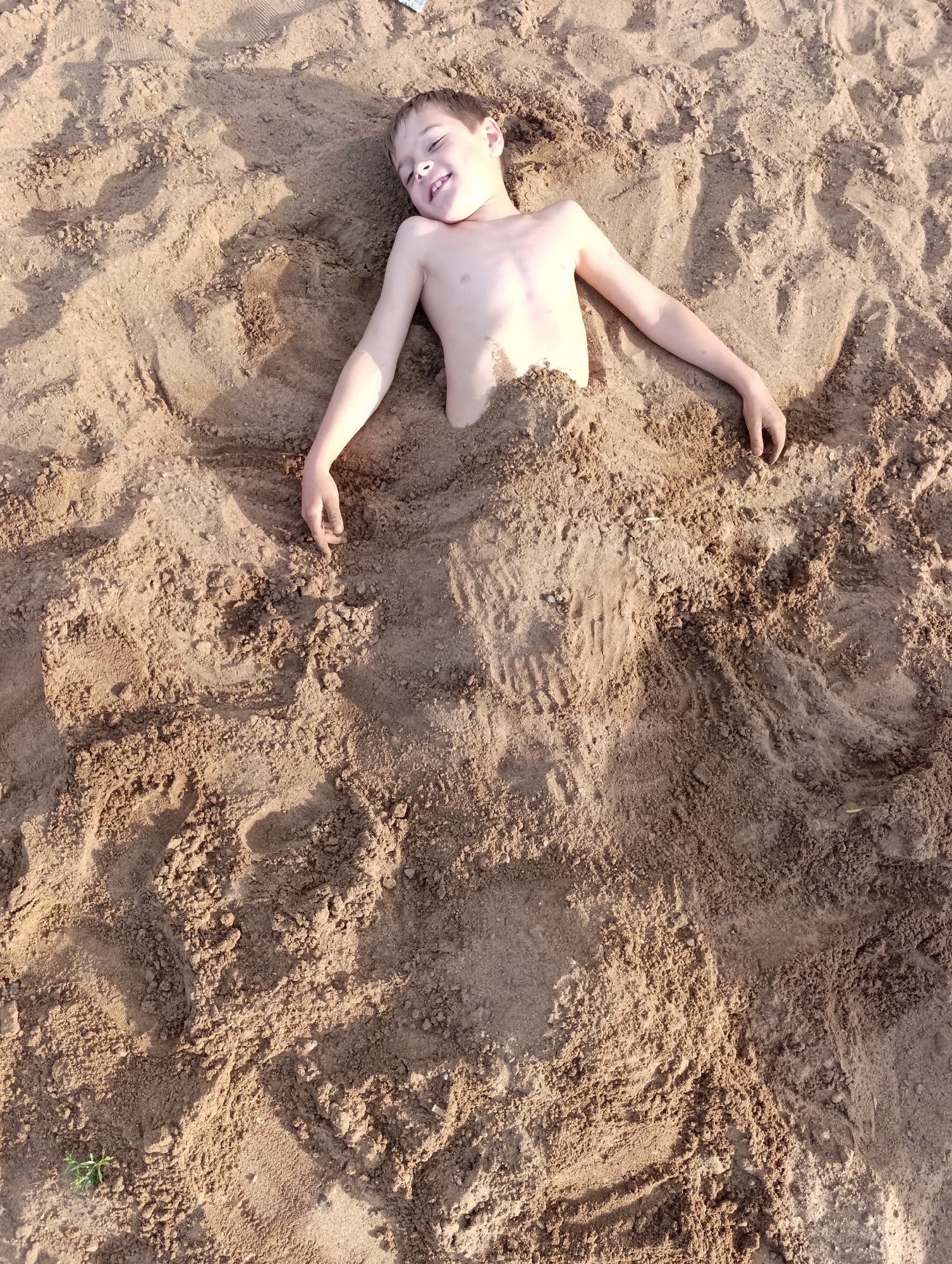 A young Caucasian boy is lying buried in sand, with only his head, shoulders, and arms exposed. He is wearing a light-colored bracelet on his left wrist and appears to be smiling or laughing, with his eyes closed. The sand is light brown and appears to be on a beach or in a sandbox. His arms are extended to his sides. The boy is positioned in the upper center of the frame. The background consists of more sand, with some shadows and indentations, suggesting other footprints or disturbances in the sand. The image is taken from a high angle looking down. The location is not specified in the image.
