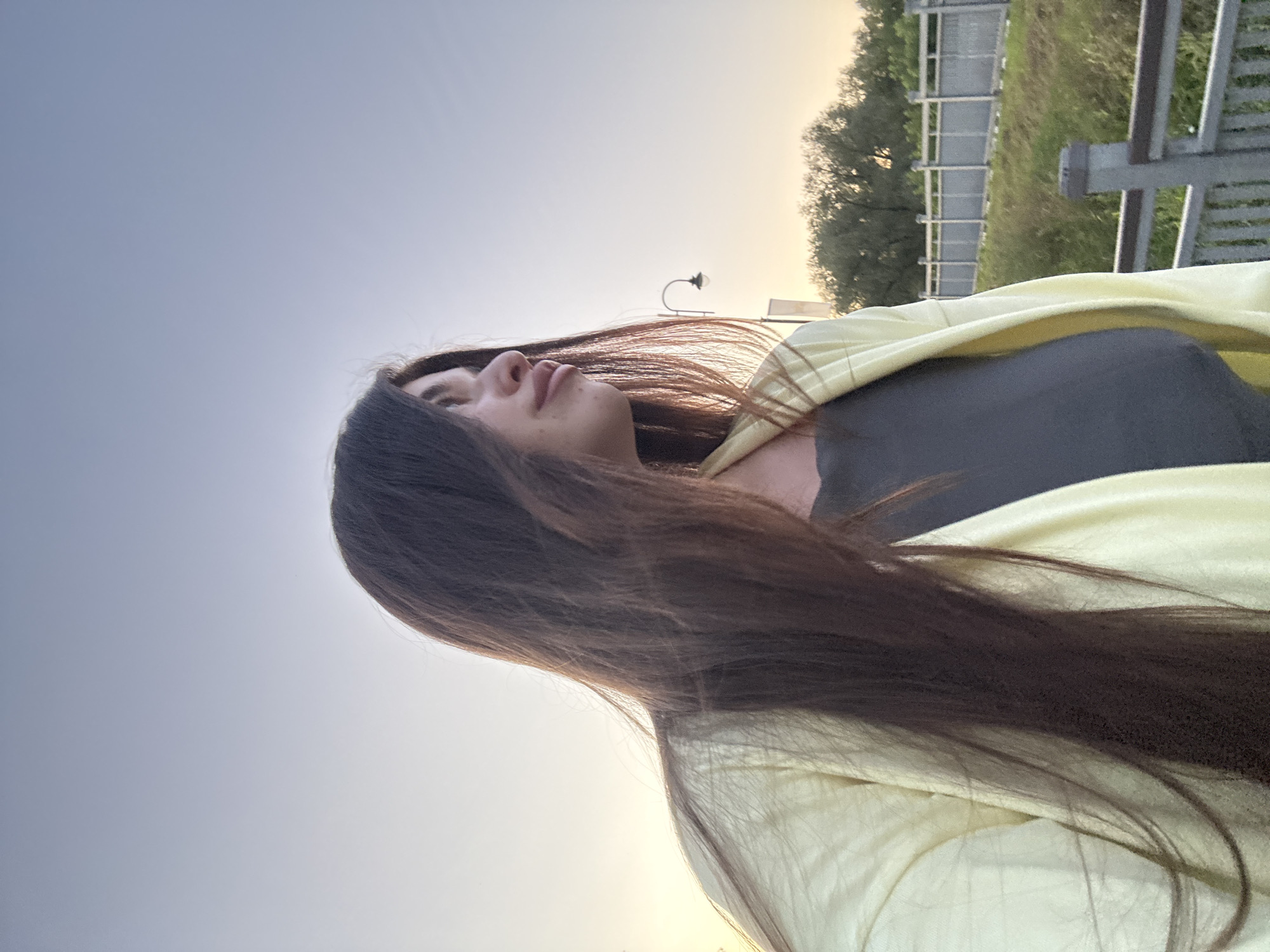 A young woman with long, dark hair is pictured in an outdoor setting, likely in the city of Klin, Russia, as suggested by the background. She is wearing a light yellow jacket over a dark grey top. The woman is looking off to the right side of the frame, with her profile visible. In the background, a metal railing and fence are present, along with trees and a lamppost, suggesting a park or promenade area. The sky is a soft, gradient blue, indicating it is either early morning or late evening. The lighting is gentle, casting a warm glow.