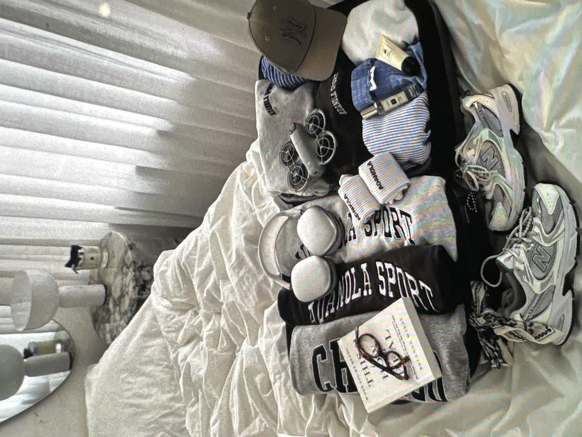 A disorganized collection of personal items is scattered on a wrinkled white bed sheet. The items include clothes (sweatshirts, striped shirts, socks, hat), accessories (headphones, glasses), a book, toiletries, a drone, and a pair of white and silver athletic shoes. The room is lit by natural light coming through closed blinds. A decorative vanity mirror is visible in the background. The location is not identifiable as being in Yerevan, Armenia.