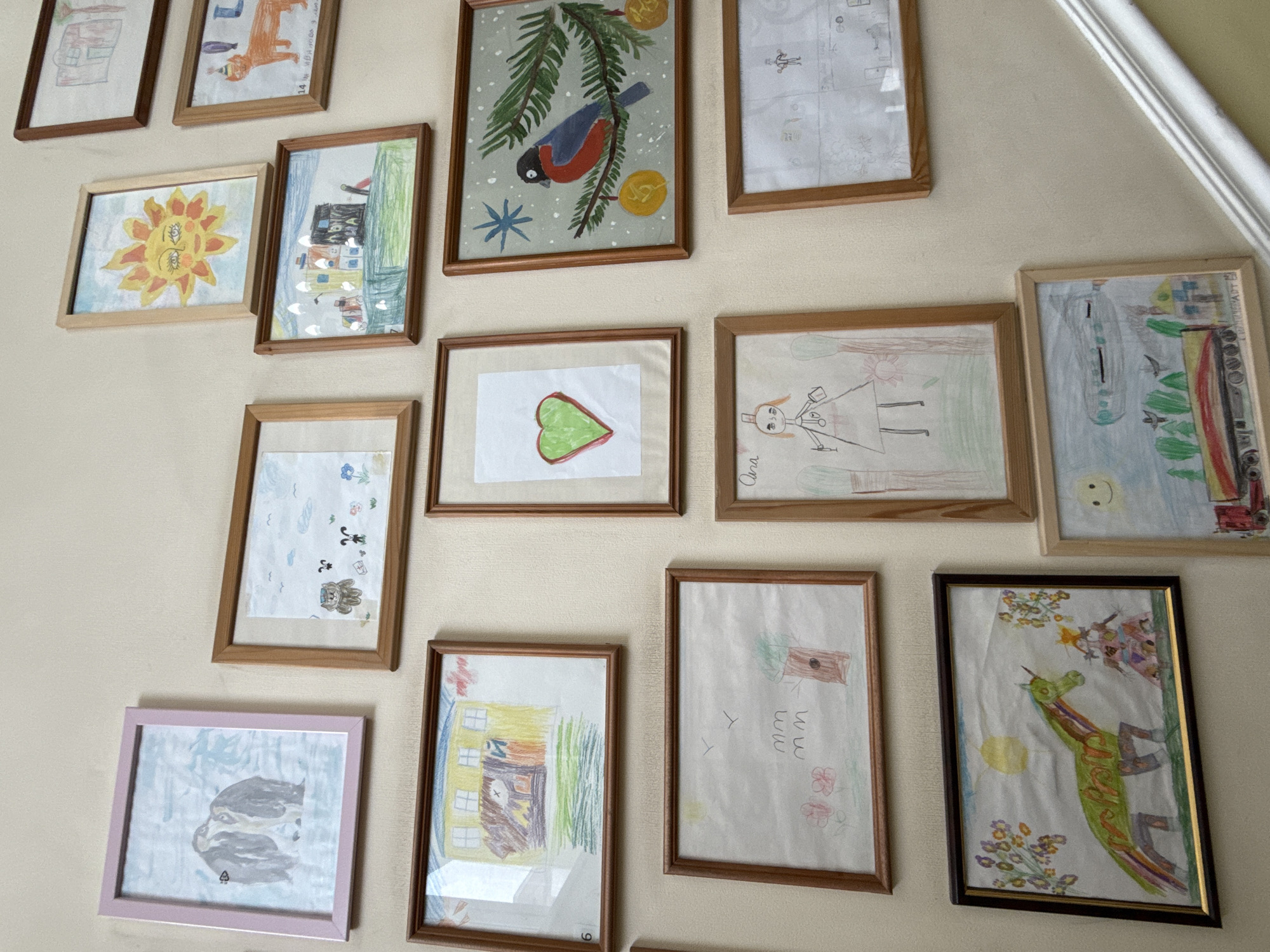 The image shows a wall decorated with numerous framed children's drawings. The drawings depict a variety of subjects including animals (penguins, a bullfinch, a cat, a horse), a sun, houses, a city skyline, a heart, and a figure that appears to be a princess. The frames are mostly wooden, with one pink frame. The wall is a light beige color. The lower right portion of the image shows a white painted staircase railing. The scene appears to be indoors, likely in a home or school. Without further information or context, it is not possible to determine the specific location in Saint Petersburg, Russia.