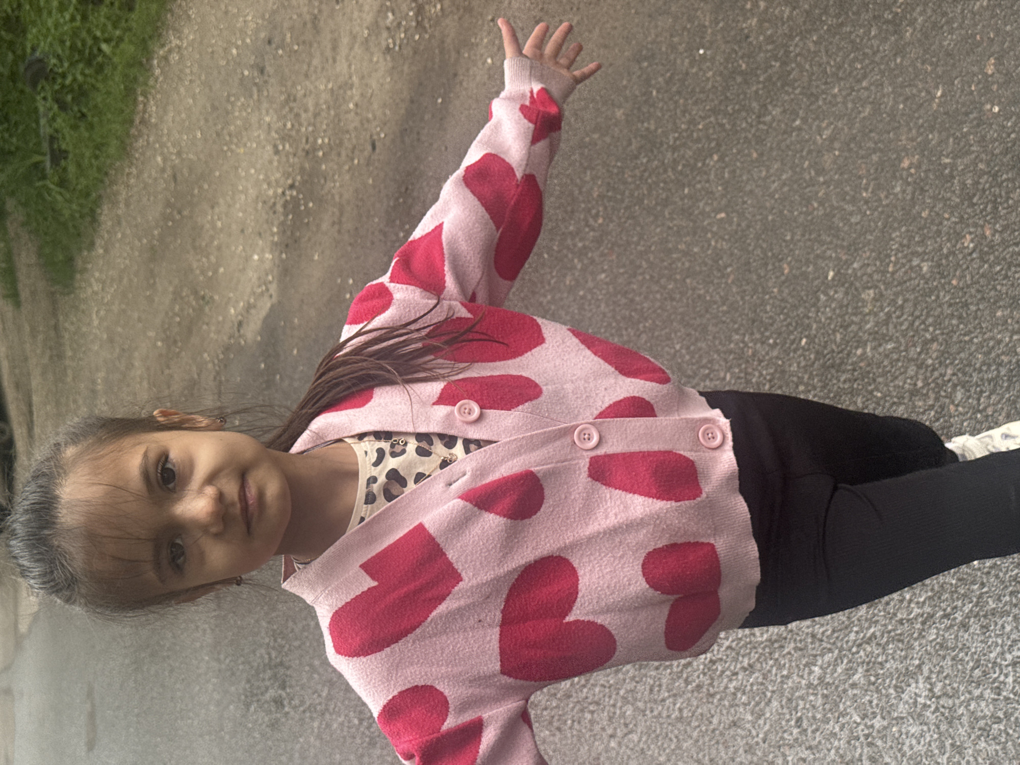 A young girl is captured in an outdoor setting, standing on what appears to be a paved, gray surface like asphalt or concrete. The image is oriented vertically, with the girl positioned upright relative to the frame. She is looking directly at the camera with a soft, friendly expression, and her dark hair frames her face.
She is wearing a light pink cardigan with numerous prominent red heart patterns. Beneath the cardigan, a cream or light-colored top with a subtle leopard print design is visible. She also wears dark, likely black, leggings and white sneakers. Her right arm is slightly raised with her palm open and fingers spread.
To the left side of the frame, there is a band of green foliage, suggesting grass or bushes, indicating a natural element in the otherwise urban or paved environment. The lighting appears soft and diffused, possibly indicating an overcast day or a shaded area, as there are no harsh shadows. No text is visible in the image. The overall impression is one of youthful cheerfulness.