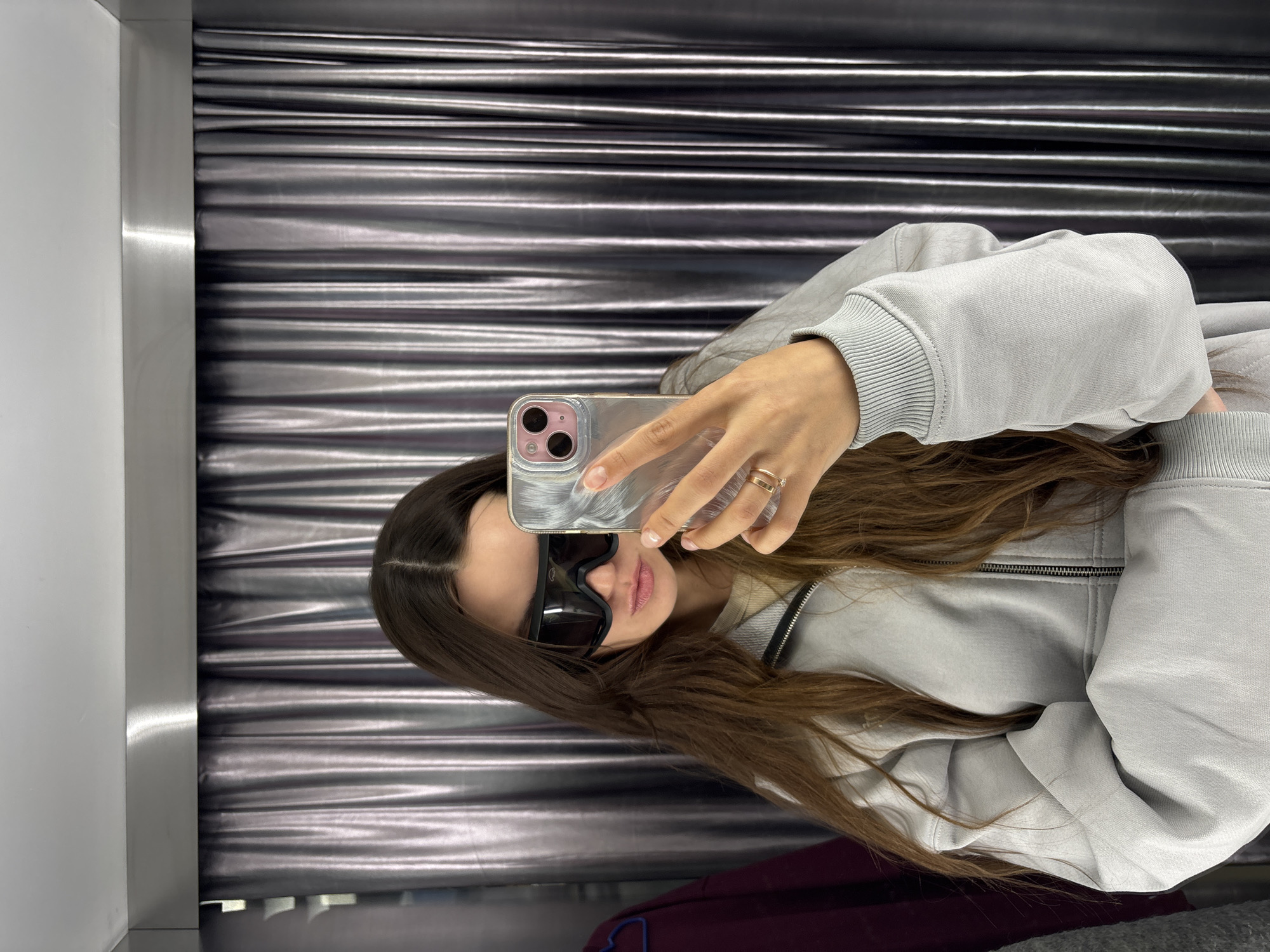 A woman wearing sunglasses and a light grey zip-up hoodie is taking a selfie with a smartphone in a dressing room. The walls of the dressing room consist of a metallic silver corrugated surface on one side and a white, non-reflective surface on the other. The location is a clothing store, most likely in Krasnoyarsk, Russia.