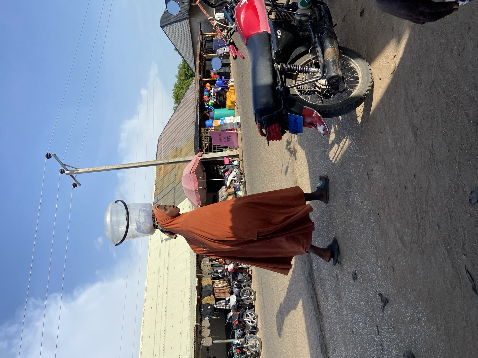 A woman walks along a road in Mararaba, Nigeria, wearing a long brown dress and carrying a clear plastic bucket on her head. She is walking near a building with a corrugated metal roof. Outside the building are various motorcycles and other goods for sale. A motorcycle is parked on the road next to her. Overhead are power lines against a clear blue sky.