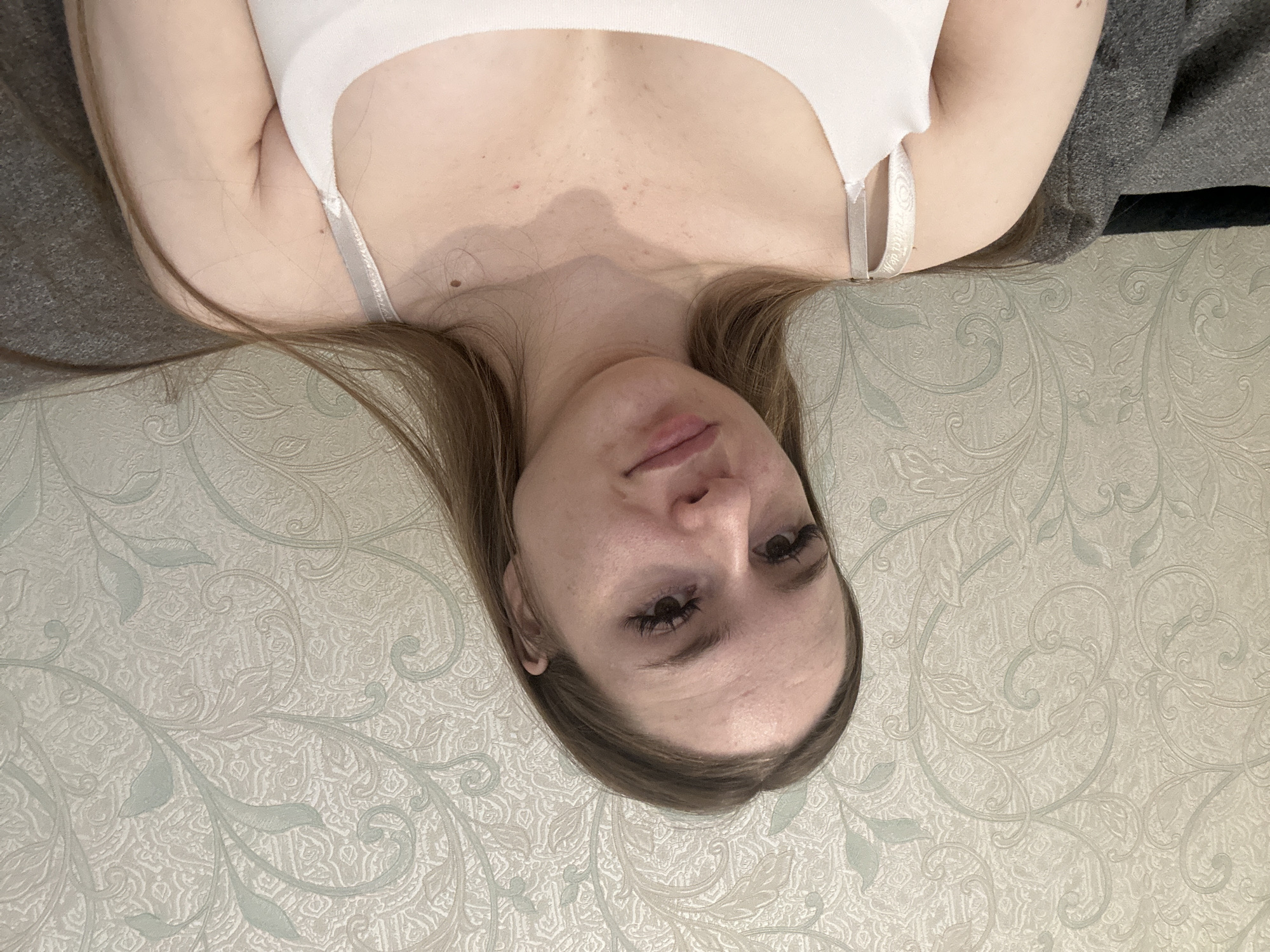 An image, rotated 90 degrees clockwise, depicts a person, likely a woman, lying down in an indoor setting. She has long, light brown hair and fair skin, and is wearing a light-colored, possibly white, sleeveless top or bralette. Her head is positioned towards the bottom of the frame, with her body extending upwards.
The person looks directly at the camera with a neutral to slightly relaxed expression. Visible details on her face include small blemishes on her forehead and distinct, possibly enhanced, eyelashes. Additional small red spots are noticeable on her chest. On the right strap of her top, the text "wild joy" is visible in a stylized, possibly circular, font on a light grey band.
The background primarily features patterned wallpaper with light-colored, intricate floral or vine motifs, possibly in cream or very light green hues. To her left and right sides (as she lies), there are fabrics; a light grey one on her left and a darker grey one on her right, possibly part of clothing or bedding. The scene conveys a casual, relaxed indoor moment. The time of day and weather are not discernible.
