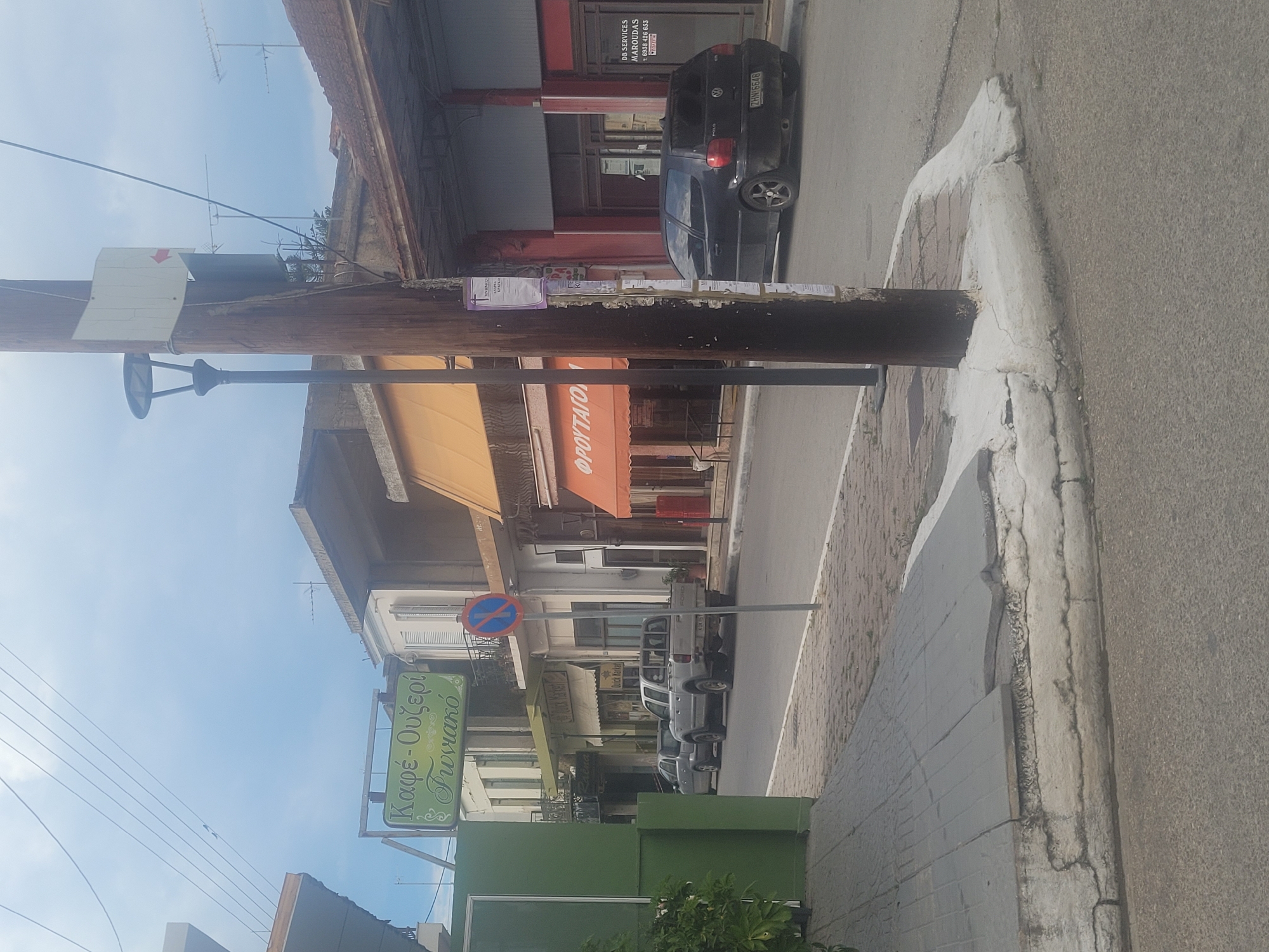 The image shows a street scene in an urban area, possibly in Greece based on the signs and text visible. A wooden utility pole stands prominently in the foreground, with a street lamp attached. The sign "Καφέ - Ουζερί Γωνιακό" is visible on the left, indicating a café or eatery. A blue "no parking" sign is also seen nearby.
Several vehicles are parked along the street, including a dark-colored car with the license plate "ZHN 6646." A sign for "DB SERVICES MAROUDAS" is partially visible. The buildings along the street have awnings, with one awning displaying the word "ΦΡΟΥΤΑΓΟ" (fruit). The sidewalk and curb are cracked and show signs of wear. The street is asphalt and appears to be moderately busy with parked cars. The sky is visible in the background, suggesting it's daytime.