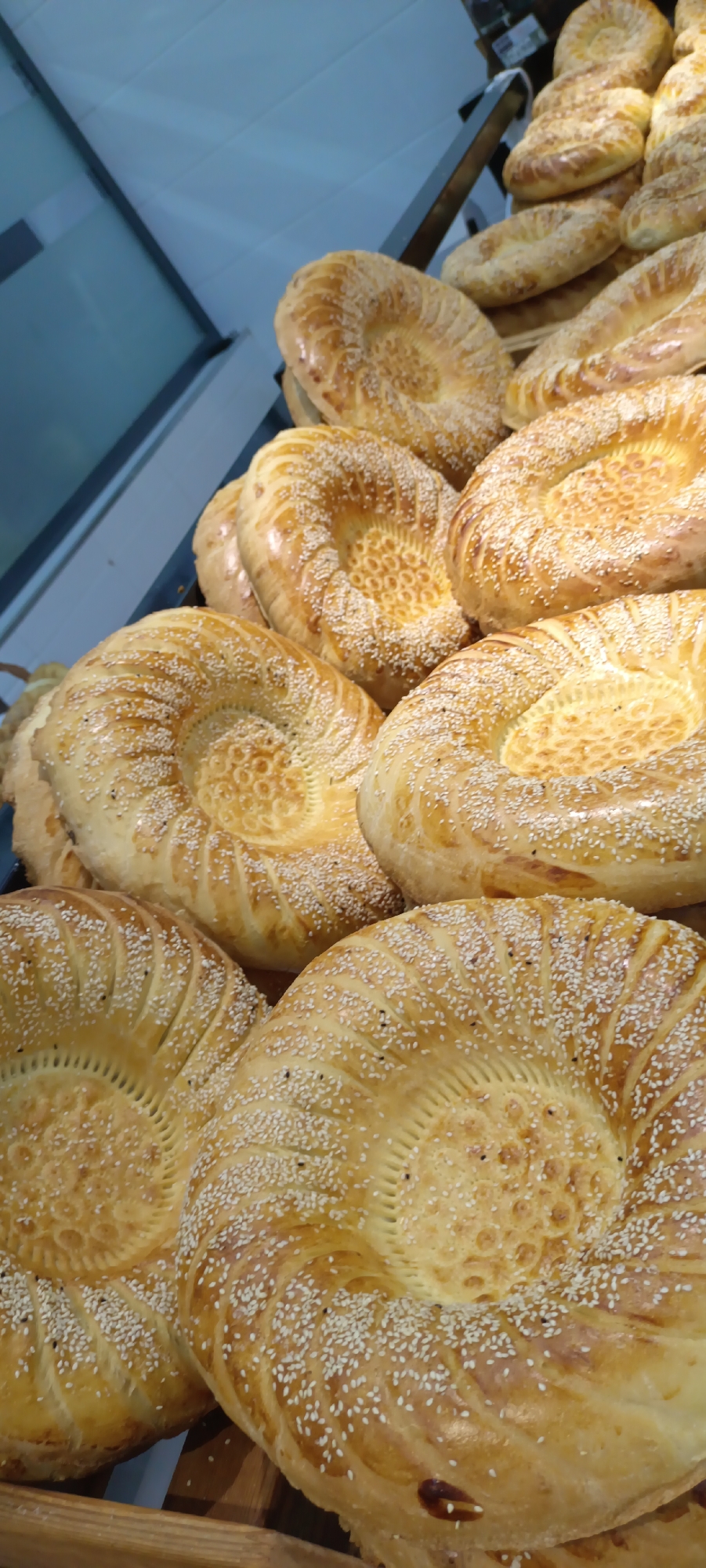 This media file shows a close-up view of several rounds of freshly baked bread, likely flatbreads, arranged on a shelf or in a display. The breads are golden brown with a slightly glossy crust, sprinkled generously with white sesame seeds. The center of each bread features an intricate pattern, created by pressing into the dough, with a central indentation. Some of the breads show a textured surface with radial lines leading to the center. The lighting suggests an indoor setting, possibly a bakery or market stall. The focus is entirely on the bread, highlighting its texture and toppings. There are no people, animals, or any other objects clearly visible besides the bread and the shelving. The context provided indicates this scene is in Fergana City, Uzbekistan, which is known for its traditional baked goods. The image is likely taken during daytime given the brightness of the illumination.