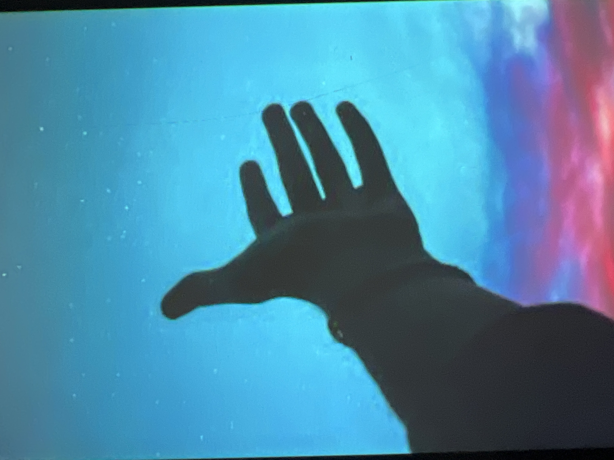 The image shows a close-up, silhouette view of a human hand outstretched towards a vibrant, abstract background. The hand, rendered in dark shadow, appears to be reaching upwards and slightly to the left. The fingers are spread, with the index and middle fingers more prominent and extended.
The background is a gradient of colors, predominantly a bright, luminous blue, interspersed with swirling clouds of pink and red. This colorful backdrop suggests a scene that is either artistic, abstract, or possibly depicting a celestial event or nebula. The blue area is speckled with small, bright dots, which could represent stars or other light sources.
There are no identifiable people, objects, or specific location cues in the scene. The lighting creates a dramatic silhouette of the hand against the colorful, luminous background, giving a sense of awe, exploration, or reaching for something unattainable. The overall mood is abstract and possibly emotive, hinting at themes of aspiration or wonder. There is no visible text in the image. The time of day and weather are not discernible from the abstract background.