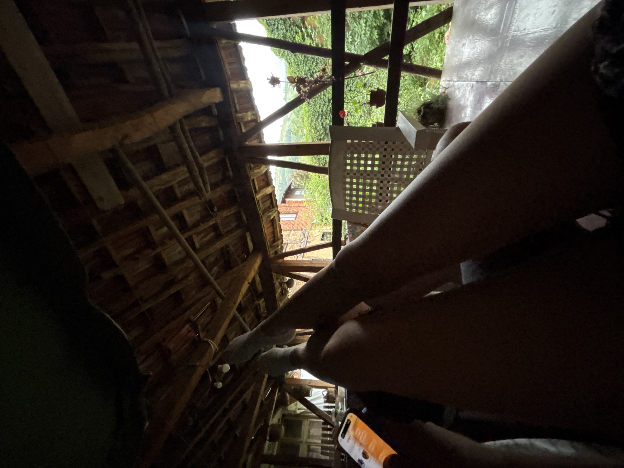 The image shows a person's legs and feet in the foreground, with their phone in their left hand. The legs are crossed and appear to be resting on a surface. The scene appears to be on a covered balcony or porch, with a wooden roof and beams visible. Beyond the balcony, there is a view of lush greenery, rolling hills, and a brick building. The location is identified as Shipkovo, Bulgaria. The overall atmosphere is relaxed and informal, suggesting a moment of leisure.