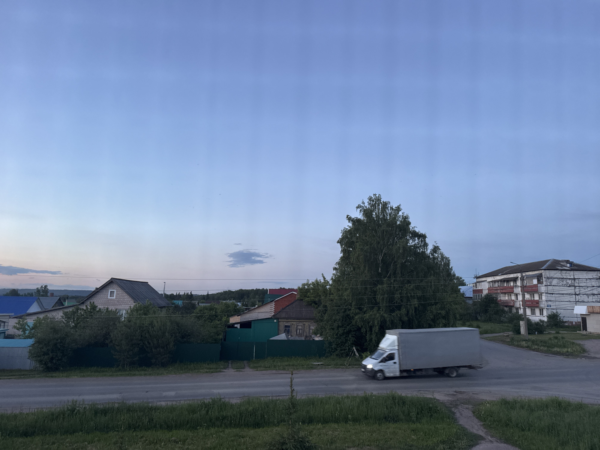 The image depicts a tranquil street scene in Mozhga, Russia, captured during the late afternoon or early evening under a light blue, partly cloudy sky. The lighting is soft and diffused, with a single distinct white cloud visible in the upper center.
In the foreground, green grass and dirt verges line an asphalt road. A white cargo van with a grey cargo box, characteristic of light commercial vehicles, is in motion from right to left across the road, its lights faintly illuminated.
The background features a mix of residential buildings and abundant greenery. On the left, several houses are visible, including one with a dark grey roof, light-colored walls, and a small white window in its gable, surrounded by bushes and green fences. Further to the right, a large, dense deciduous tree dominates the mid-ground. Beyond the tree, a multi-story apartment building with white and grey facades and balconies is visible, showing signs of wear or ongoing repair with scaffolding-like structures on its right side. Power lines crisscross the scene above the buildings and trees. The overall impression is of an ordinary, calm moment in a provincial town. There is no visible text in the image.