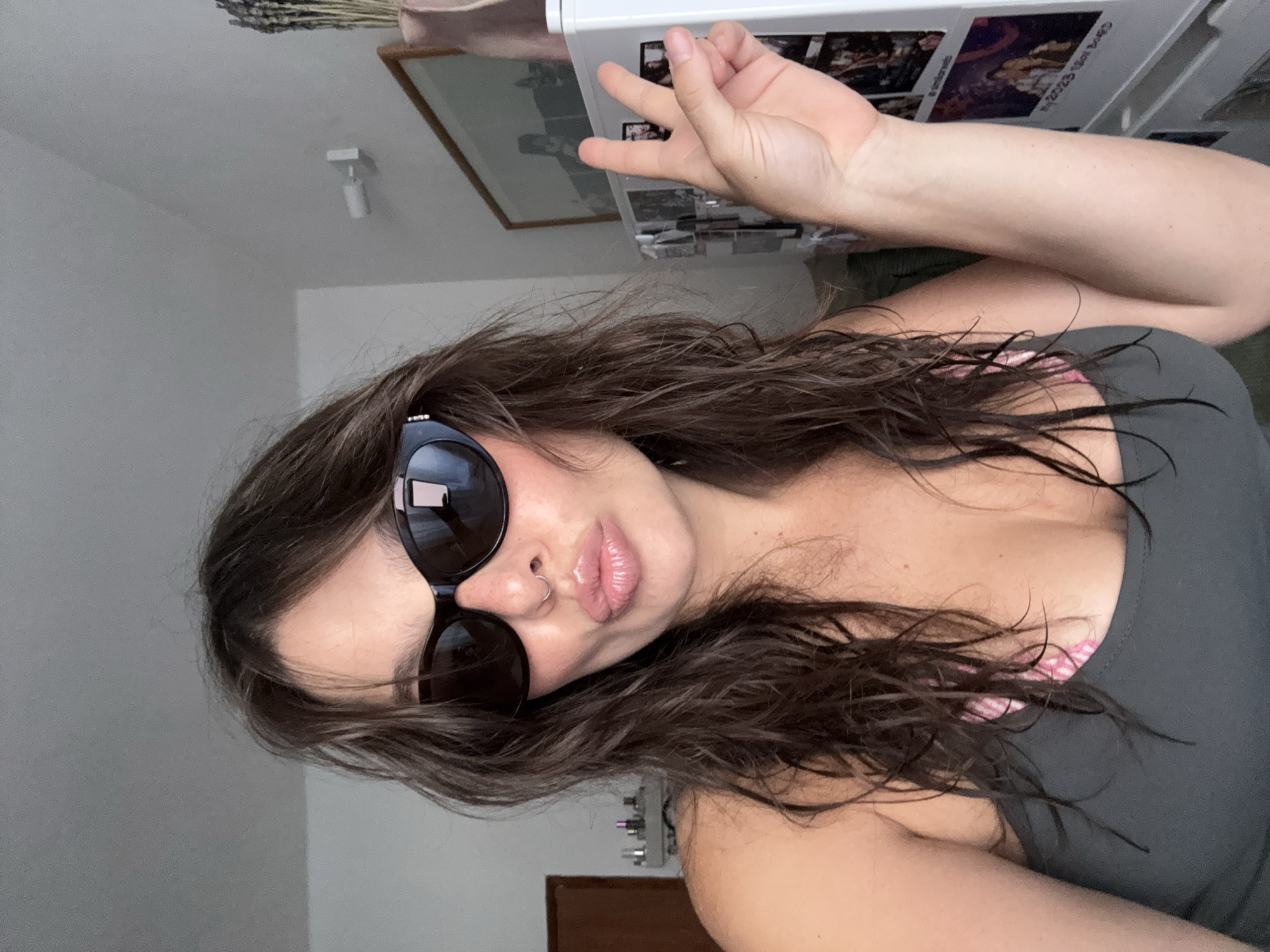 A woman with long, wavy brown hair and wearing sunglasses is facing the camera and making a hand gesture. She has a nose ring and full lips. She is wearing a gray tank top with a glimpse of pink pattern beneath. In the background, there is a picture frame on the wall and a white object with printed images. The background also includes a ceiling light, and what appears to be a small wooden door near the bottom of the frame. There is no clear evidence indicating this photo was taken in Tel-Aviv, Israel.