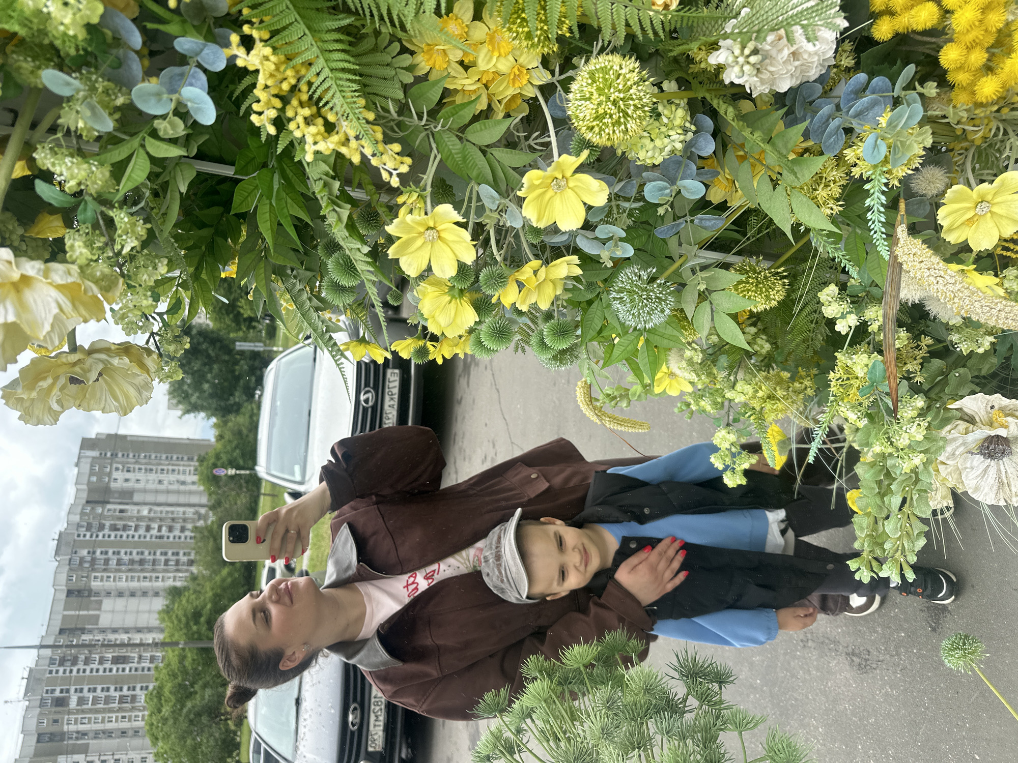 In Moscow, Russia, a woman is taking a selfie with a child in front of a large, decorated floral archway. The woman, with her hair in a bun, is wearing a brown jacket over a pink t-shirt and is holding a smartphone to capture the photo. She is smiling and looking at the phone's screen. Next to her, a young boy in a gray baseball cap, blue long-sleeved shirt, and black vest is looking towards the camera with a slight smile. The floral archway is lush with various green foliage and an abundance of yellow flowers, creating a vibrant and festive backdrop. In the background, several cars are parked, and a tall, multi-story residential building with a grid-like facade is visible, characteristic of Soviet-era architecture. The sky is overcast, suggesting a cloudy day. The scene appears to be set in an outdoor urban environment, possibly a park or a public area decorated for an event.