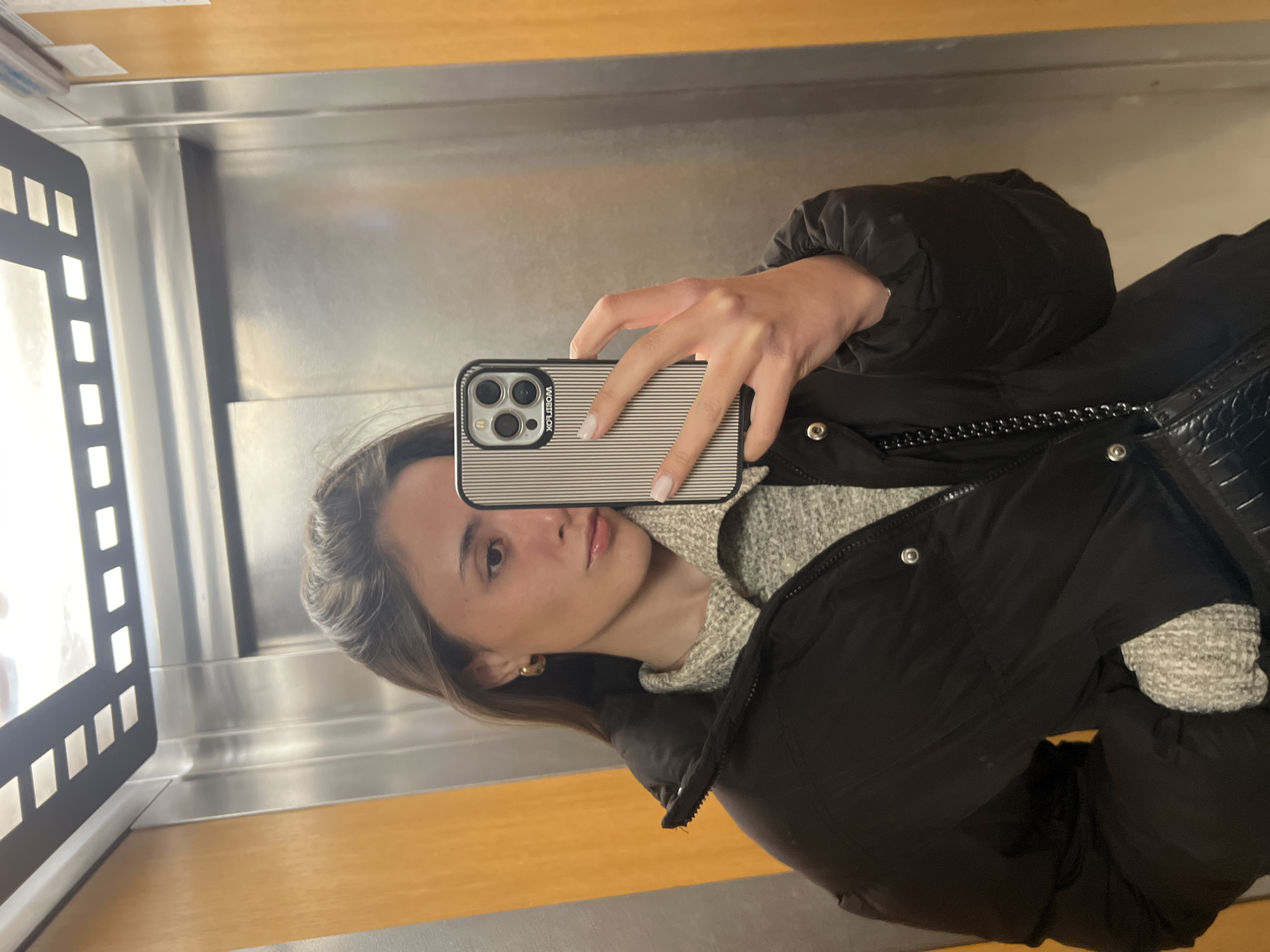 A woman is taking a selfie in an elevator. She has light brown hair and is wearing a grey sweater and black puffer jacket. Her phone is in front of her face, obscuring the lower half. The elevator has a silver interior and a wooden floor. There's a black panel to the left with rectangular cutouts. The location is Sofia, Bulgaria, though this cannot be determined from the image.