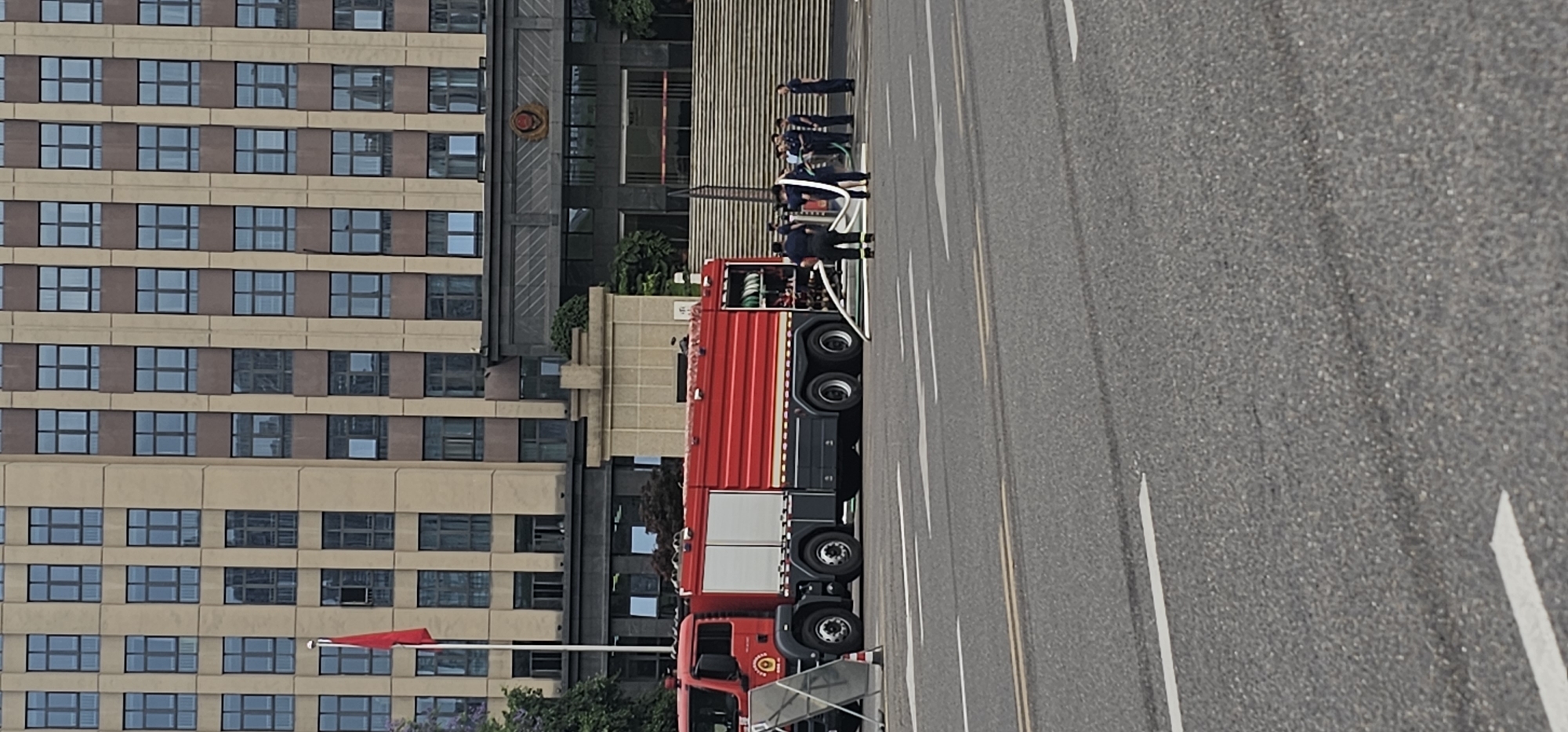 The scene depicts a fire department operation in the Jiulongpo District, China. A red fire engine is parked on an asphalt road with white lane markings. Several uniformed firefighters are standing nearby, some handling white hoses. A tall building with numerous windows is visible in the background, with a red flag displayed on a pole in front of it. The environment appears to be a city street.