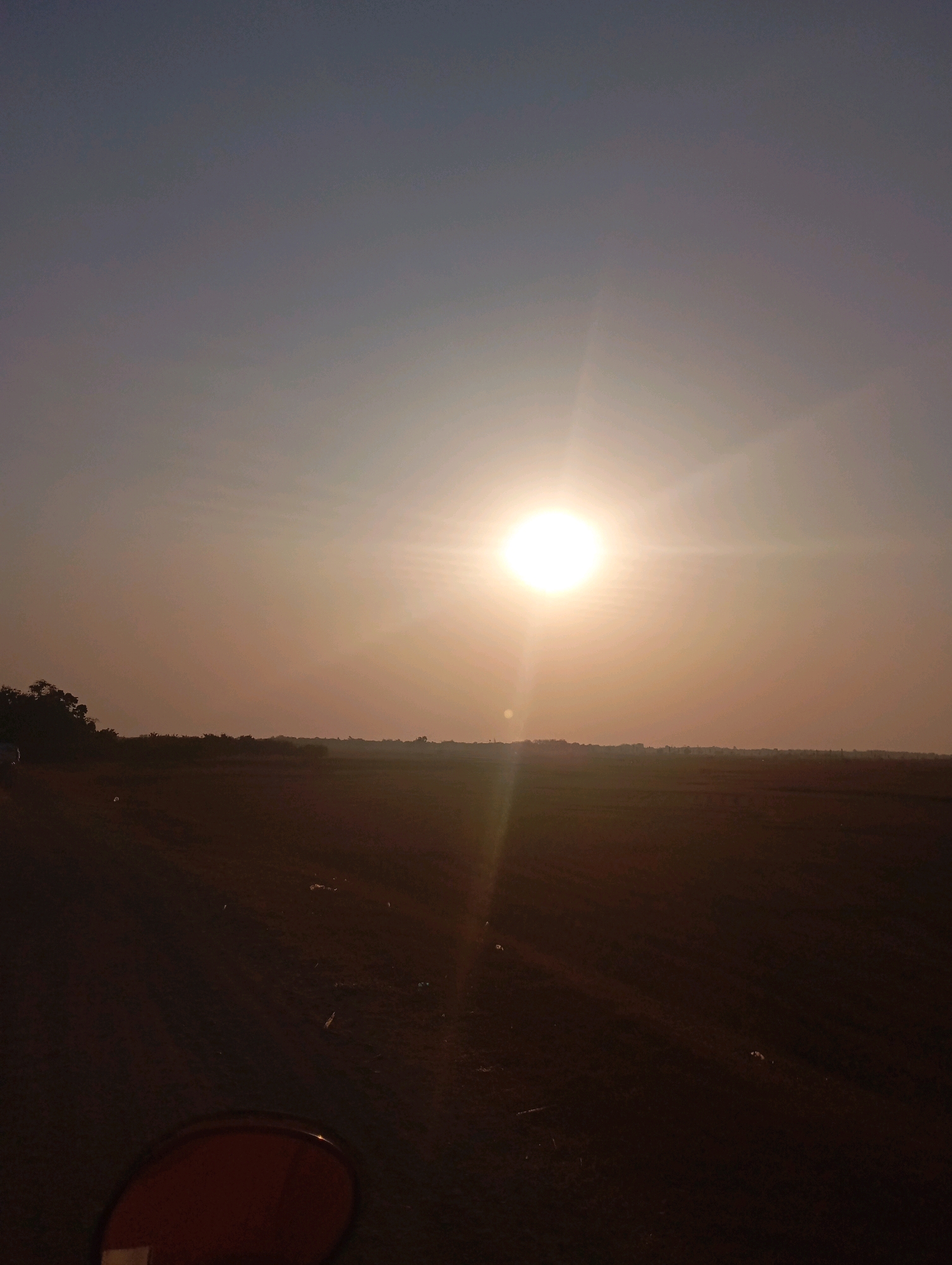 The image depicts a wide, open landscape under a hazy sky, dominated by a bright sun positioned slightly left of center, casting a warm glow and creating lens flare. The ground appears to be a field, possibly agricultural, with a dirt road or path leading from the lower left corner into the distance. A line of trees and vegetation forms a distant horizon. In the lower foreground, a circular, reddish-brown object, possibly part of a vehicle or equipment, is visible. The overall impression is one of a rural or semi-rural setting at either sunrise or sunset. The image is consistent with a location in Haurkolot, Indonesia, given the flat terrain and vegetation typically found in such areas.