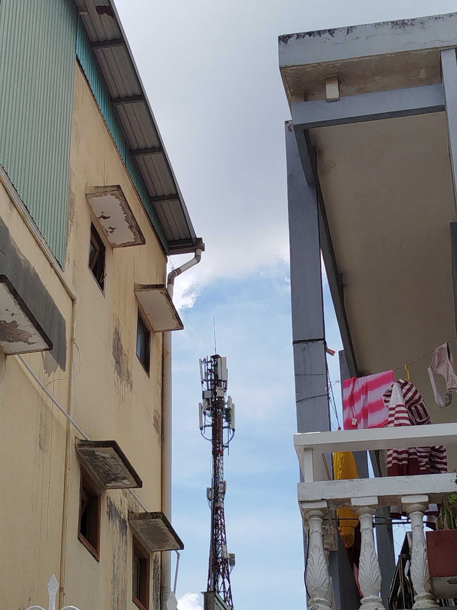 The image shows a view of buildings and a cell tower against a sky with clouds. On the left, a building with a corrugated metal roof is visible, with a yellow-painted facade and several windows. One window is open, and another has a small awning. A rain gutter runs down the side of this building. In the center, a tall cell tower with multiple antennas stands against the sky. To the right, a portion of another building is visible, with a concrete balcony and railing. Laundry, including striped fabric and undergarments, is hanging on a clothesline on the balcony. The overall scene suggests an urban or semi-urban environment.
