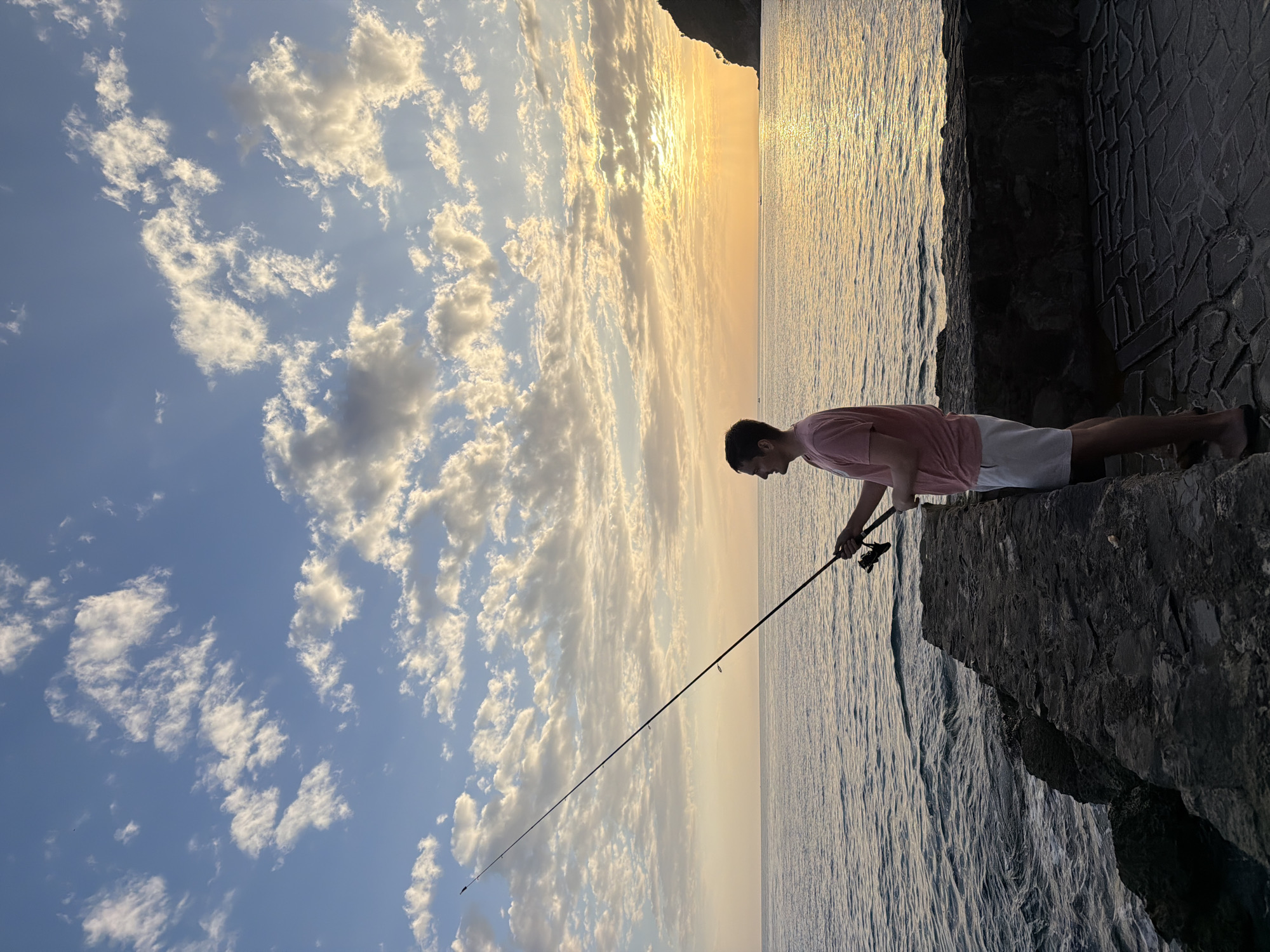 A man is fishing in Petrovac centar, Montenegro. He stands on a rocky ledge, wearing a pink t-shirt and white shorts, holding a fishing rod over the water. The sea is calm with visible ripples, and the sky is filled with clouds as the sun sets on the horizon. A part of a stone structure, possibly a wall or building, is visible behind the man.