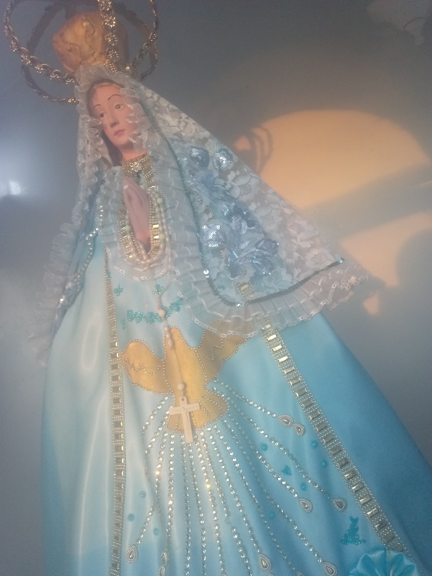 The image shows a statue of the Virgin Mary in Juan Griego, Venezuela. She is wearing a light blue robe adorned with gold trim, a golden dove emblem, and strands of silver beads and teardrop embellishments. Her head is covered with a white lace veil. She wears a golden crown. The statue is against a light-colored wall, with an area of light and shadow behind it.
