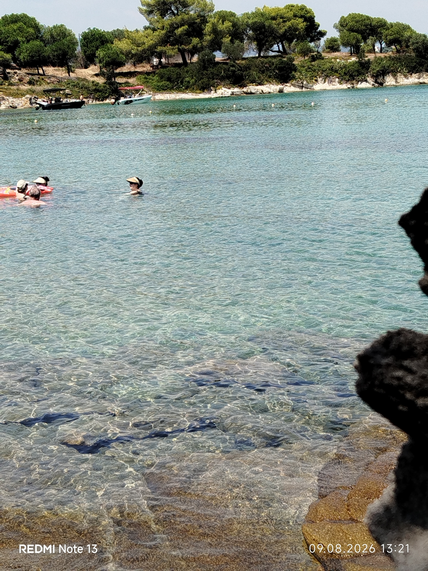 The image shows a clear, shallow body of water with a rocky shoreline and lush green trees in the background. In the water, there are several people swimming or relaxing. On the left, three individuals are partially submerged, with one person wearing a hat and another appearing to be on an inflatable object. To the right of them, a person wearing a sun visor is also in the water. Two boats are visible further out in the water, near the shore, with one appearing to be a small motorboat and the other a recreational boat. The water is transparent, revealing the rocky seabed below. The overall scene depicts a serene coastal environment, likely a beach or cove in Sithonia Municipal Unit, Greece, on a sunny day.