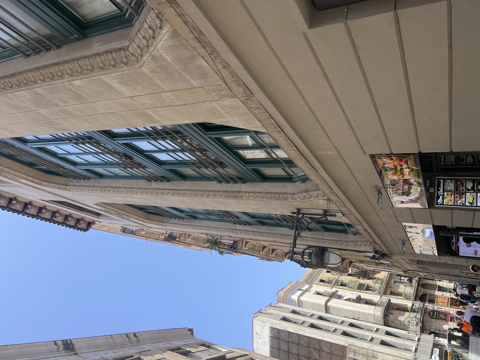 The image shows a street view in Barcelona, Spain, with buildings on either side of a narrow street. The building on the right is a tan-colored structure with ornate architectural details, including decorative moldings around the windows. The windows are large and have dark green frames, with metal grilles on the outside. The building on the left is a lighter colored building with balconies and a more modern appearance.
At the bottom of the image, there are storefronts with menus and advertisements for restaurants. One sign reads "Mimi Restaurante" and displays various dishes. People are walking on the sidewalk, some are standing and looking at menus. A white truck is parked on the left side of the street. The sky is clear and blue. The perspective is looking up from street level, emphasizing the height and architecture of the buildings.