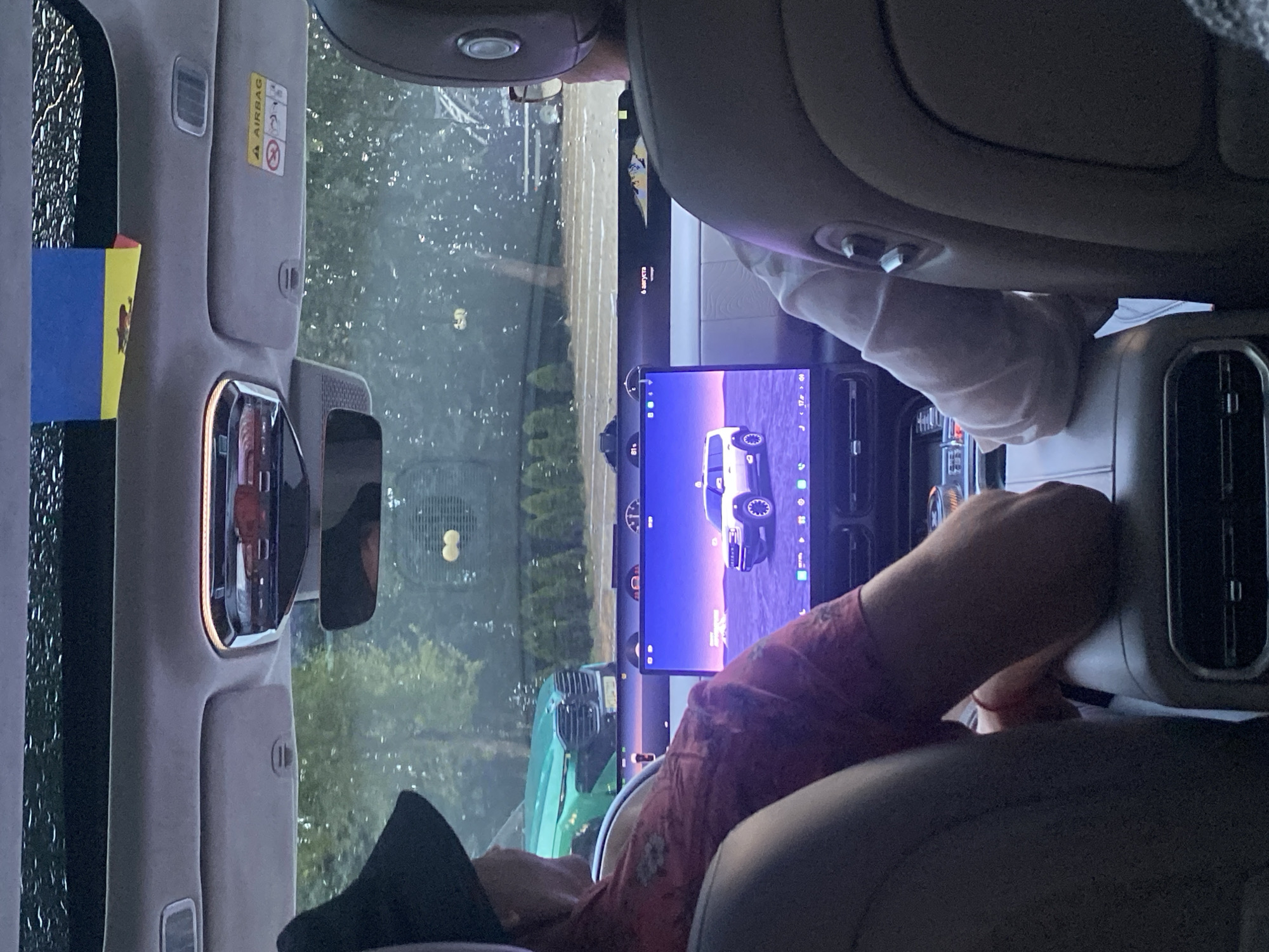 In the foreground, two individuals are seated inside a vehicle, viewed from the rear passenger seats. The person on the left wears a dark hat and a red patterned shirt, leaning forward with their left arm resting on the center console. The person on the right wears glasses and a light-colored long-sleeved shirt, also with their arm resting on the center console.
The vehicle's interior is visible, including a large touchscreen display in the center dashboard showing an image of a white SUV. Above the windshield, a Ukrainian flag is affixed to the car's interior. The exterior view through the windshield shows a rainy day with trees and other vehicles, including a green SUV.
The image is dated August 6th, as indicated by text on the dashboard display. While the location of Ustia, Moldova is mentioned, there are no specific visual cues within the image that definitively confirm this location. The scene depicts occupants of a vehicle during transit.