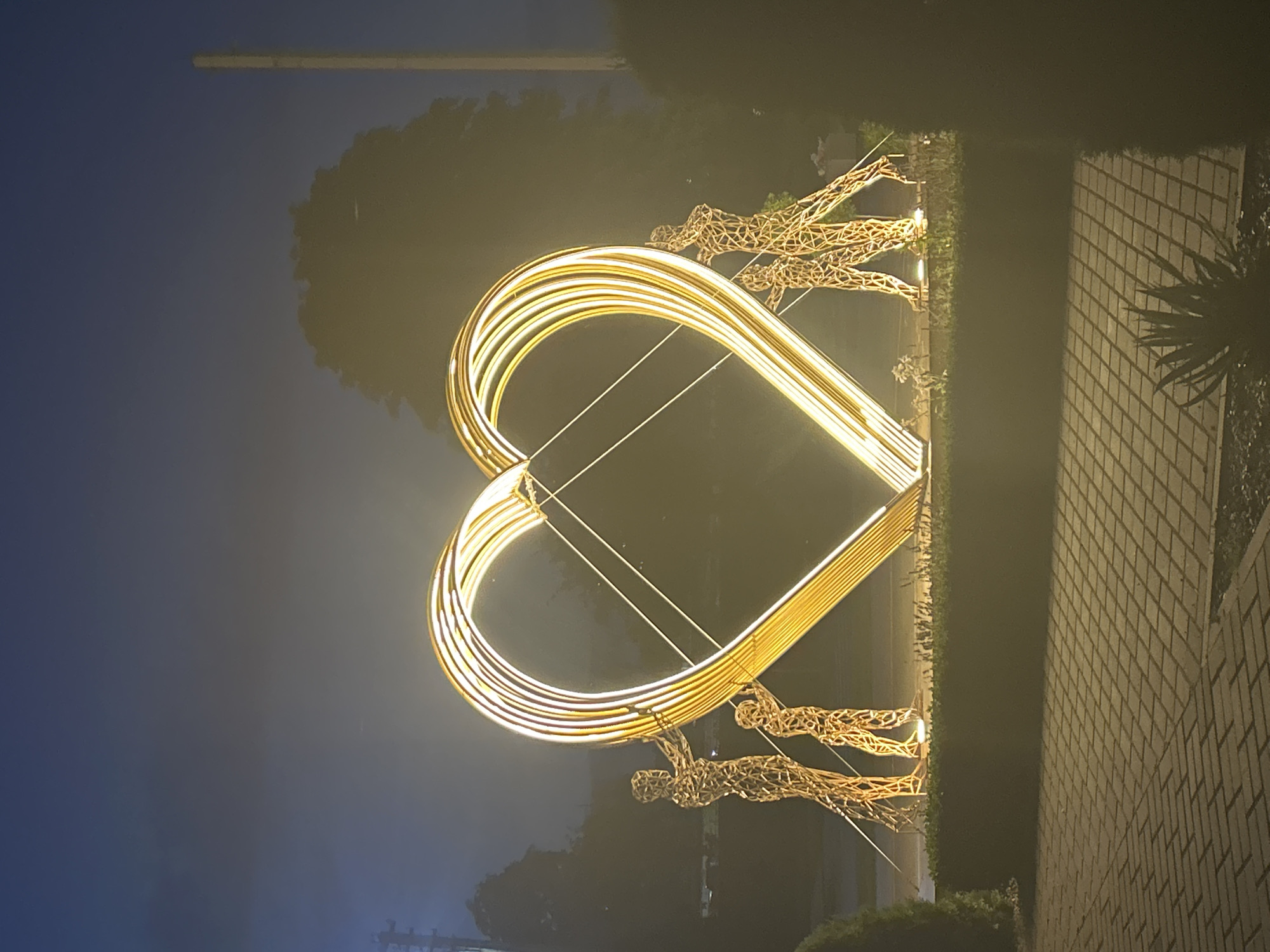 The image depicts an illuminated heart-shaped sculpture at night, situated in the city of Brest, Belarus. Four wireframe human figures are positioned around the base of the sculpture, appearing to hold or support it. Two figures are on the left side of the heart, one taller than the other, and two figures are on the right side, also with a size difference. The sculpture itself is made of golden, glowing, parallel lines that form the heart shape, with thin wires stretched across the interior. The ground in front of the sculpture is paved with brickwork. A dark green hedge runs along the base of the sculpture. The background is obscured by darkness and a misty or hazy atmosphere, with indistinct trees and structures visible. A tall, light-colored pole is present on the right side of the frame. The lighting from the sculpture casts a warm glow on the surrounding area.
