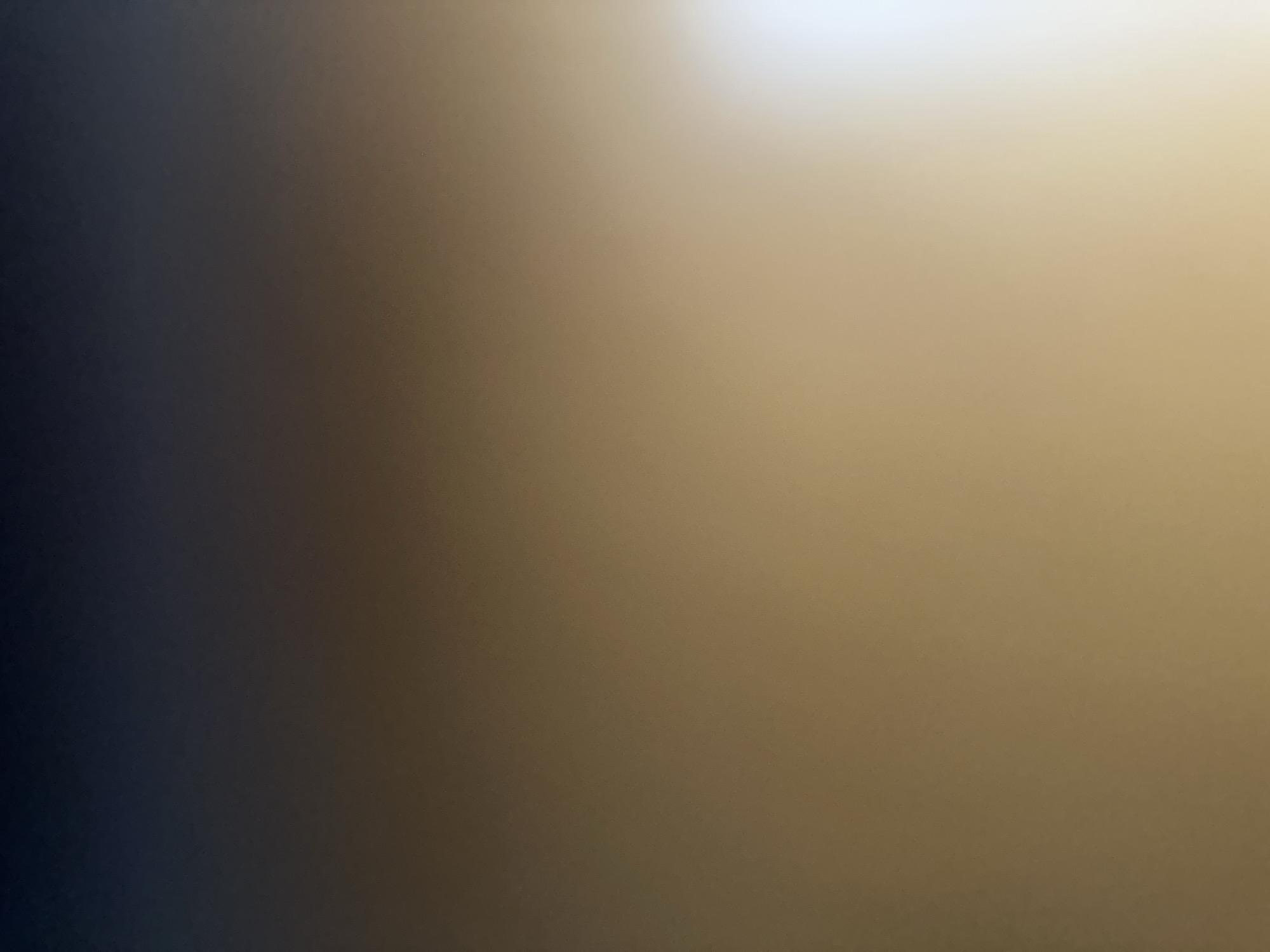 This media file is an abstract image, likely a photograph with a shallow depth of field, that exhibits a smooth gradient of colors. The left side of the image is a deep, dark blue-gray that gradually transitions to a lighter, warm beige or tan tone as it moves towards the right. The top right corner of the image is illuminated by a bright, white light source, suggesting a strong light is present in that area. There are no discernible subjects, objects, or scenes depicted in the image. The texture of the image is smooth and soft, with a subtle grain that is characteristic of photographic noise. The overall impression is one of abstract color and light, with no specific details to indicate a time of day, weather, or location. There is no visible text.