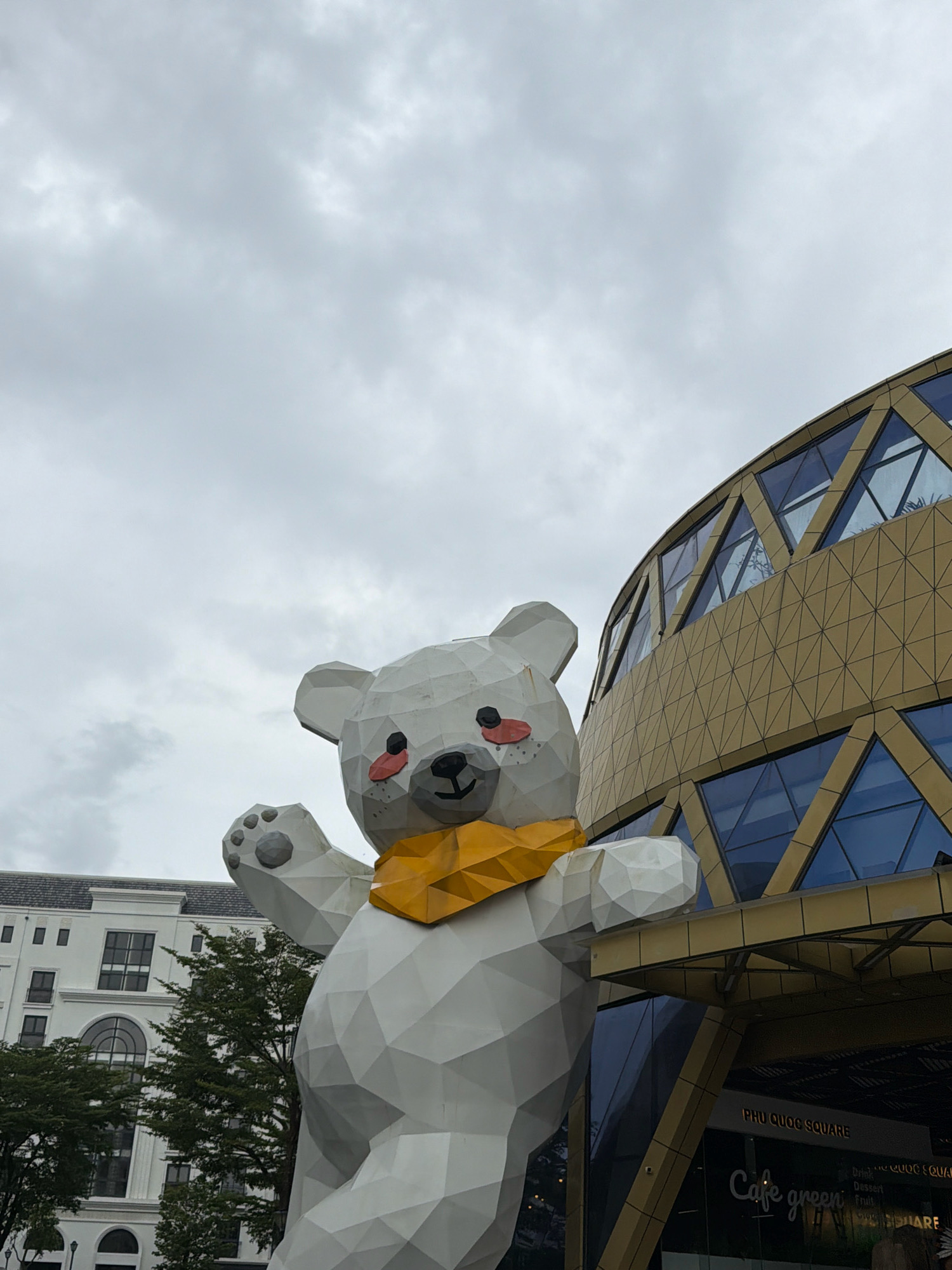 The image shows a large, white, polygonal sculpture of a bear wearing a yellow scarf, positioned in front of a modern building with a geometric, golden facade and blue glass windows. The building is identified by signage as "PHU QUOC SQUARE" and features a cafe called "Cafe green". To the left of the bear sculpture is a white building with multiple windows and arched entrances, partially obscured by green trees. The sky is overcast and grey. The scene appears to be an outdoor public space in the city of Phú Quốc, Vietnam, likely a commercial or entertainment complex. No people are visible in the image.