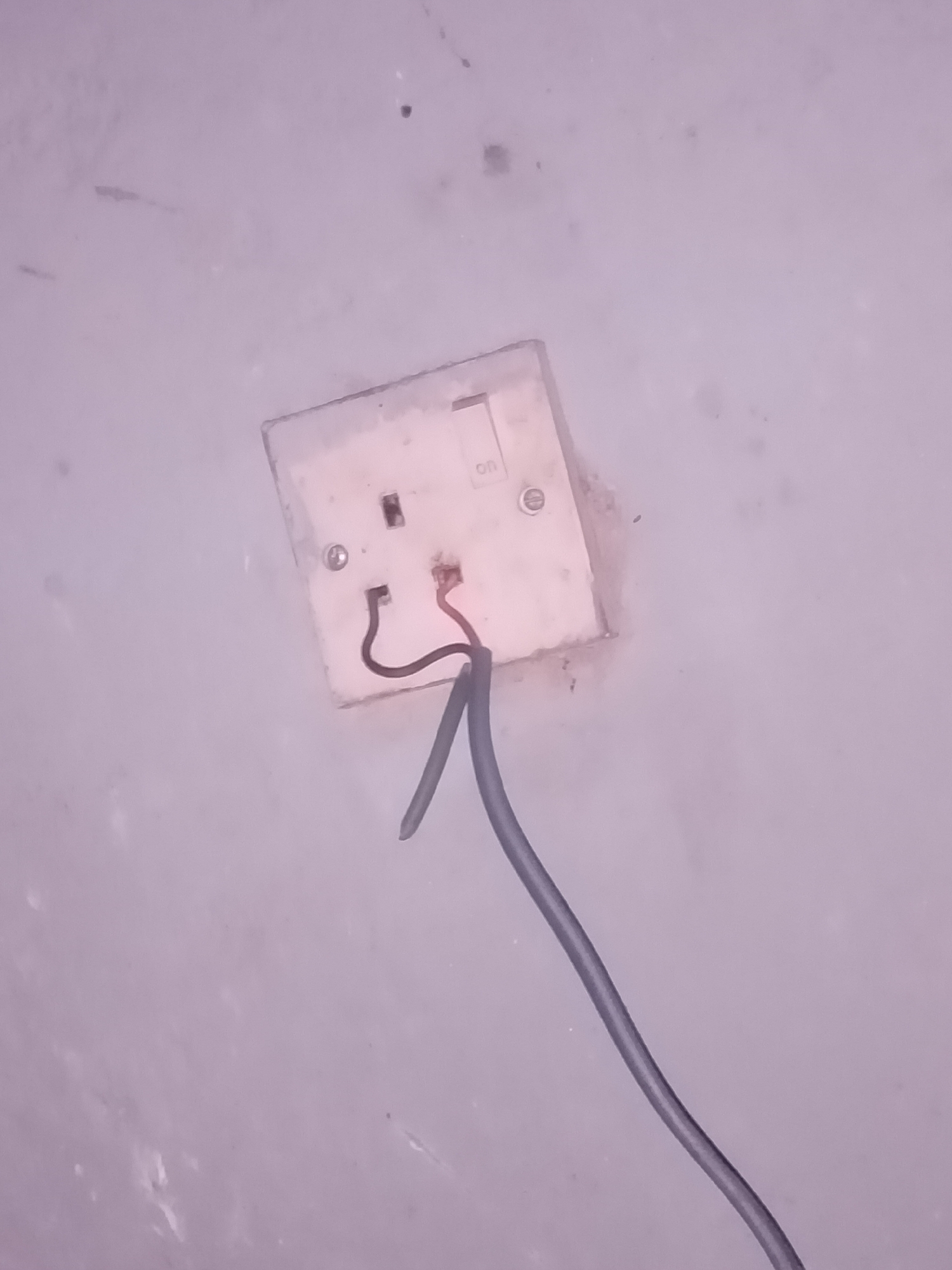 In Jalingo, Nigeria, a close-up, downward-angled shot shows a light-colored electrical outlet on a textured, light-colored wall. The outlet, which appears aged and slightly dirty, has a switch labeled "ON". Two exposed wires, one red and one dark brown, are visible emerging from the top of the outlet. A thicker, dark gray cable extends from the bottom of the outlet and curves downwards out of the frame. The lighting is dim, casting subtle shadows and highlighting the imperfections on the wall and outlet.