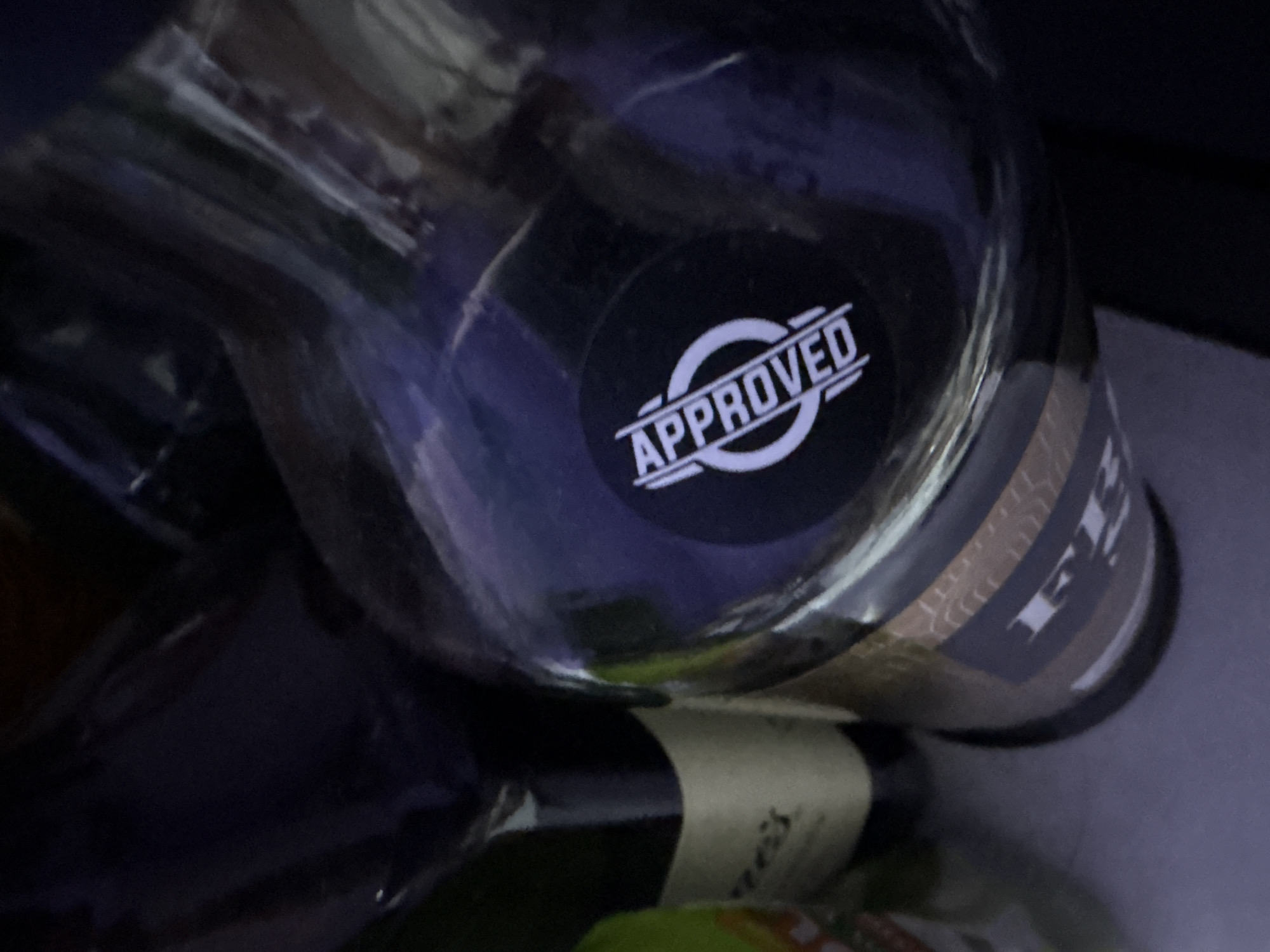 The image shows a close-up view of several bottles, likely in a dimly lit environment, possibly a store or a shelf. One bottle in the foreground, made of clear material, has a black circular sticker with the word "APPROVED" in white, stylized text. The sticker also features concentric circles. To the left, a dark bottle with a cream-colored label is visible, with some text on the label that is partially obscured and difficult to read. The overall lighting suggests the image was taken indoors. There are no people or identifiable landmarks in the image, making it impossible to determine a specific location within North Jakarta, Indonesia.