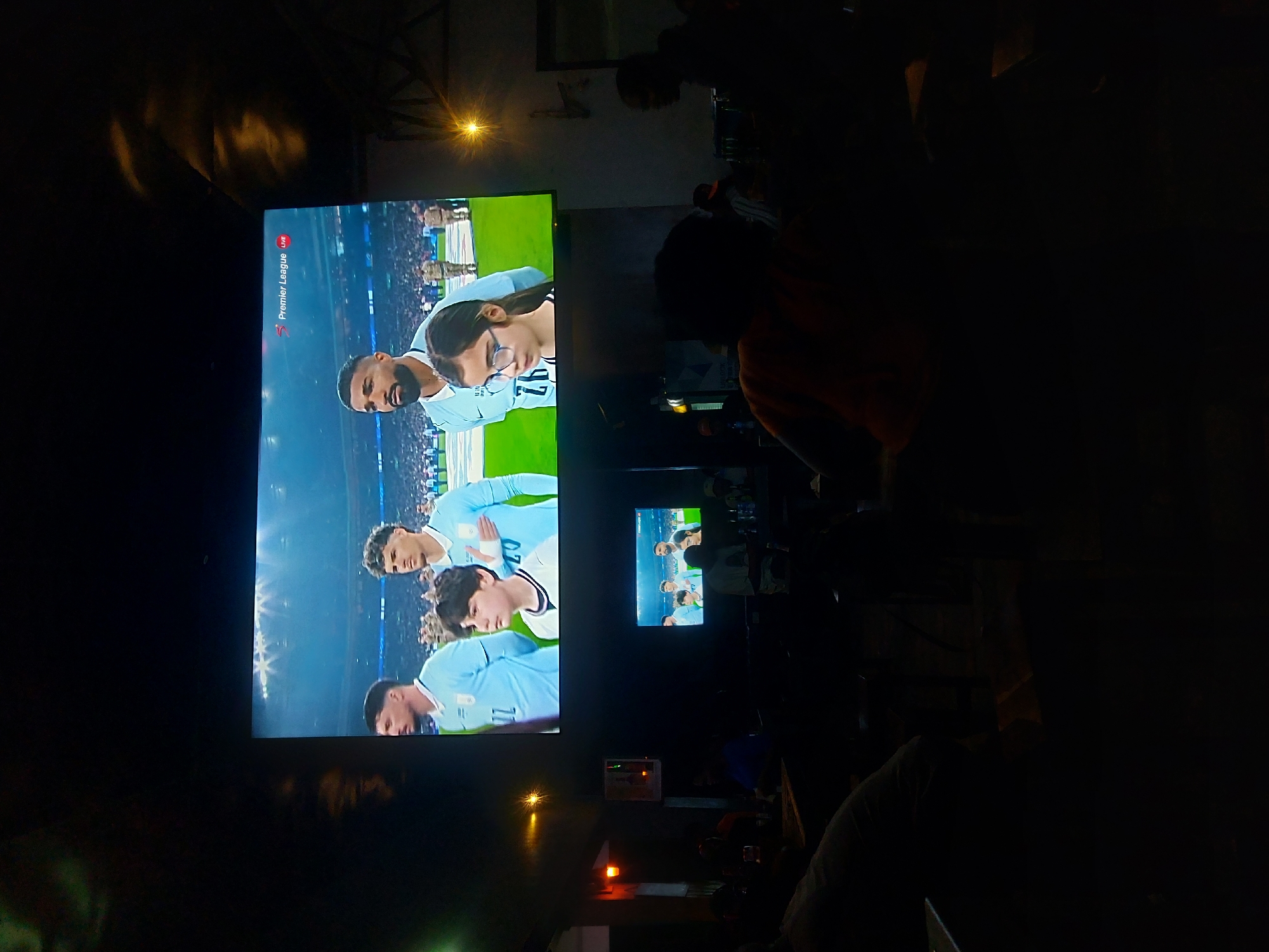 The image depicts the dimly lit interior of a public viewing establishment, likely a bar or lounge, in Dar es-Salaam, Tanzania. The specific location within the city is not ascertainable from the visual information provided.
The central focus is a large projection screen displaying a live broadcast of a Premier League football match, indicated by "Premier League LIVE" text in the top right corner. The screen shows several male football players in light blue jerseys, standing in formation on a pitch alongside a young boy and a young girl wearing glasses. A smaller television screen below the main projection replicates the same live broadcast.
In the foreground and midground, approximately six to eight individuals are visible in silhouette, seated at tables and facing the screens, suggesting they are watching the match. One person in an orange-brown shirt is prominent in the mid-right. The overall environment is dark, with ambient light emanating primarily from the screens and a few localized light sources, including a bright, star-shaped fixture on the right wall and a rectangular illuminated object with a reddish glow on the left. No explicit interactions among the audience members are discernible.