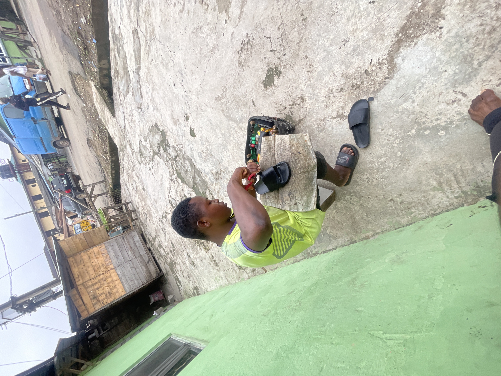 The image depicts a person in Akowonjo, Nigeria, engaged in the repair of footwear. The subject, a young man wearing a lime green t-shirt and black shorts, is seated on a wooden stool in front of a weathered, makeshift workbench. He is actively working on a black sandal, holding a tool in his right hand. A collection of footwear components, including more black sandals, are visible within a tool organizer placed on the workbench. The ground is paved with concrete, showing signs of wear and tear, with several puddles of water and debris scattered around. A vibrantly green, partially visible wall of a building stands to the left of the frame, contrasting with the neutral tones of the surrounding environment. In the background, various elements of urban life are discernible, including a wooden stall, vehicles like a blue van and a black SUV, and residential buildings. The overall atmosphere suggests a typical street scene in a bustling Nigerian city.