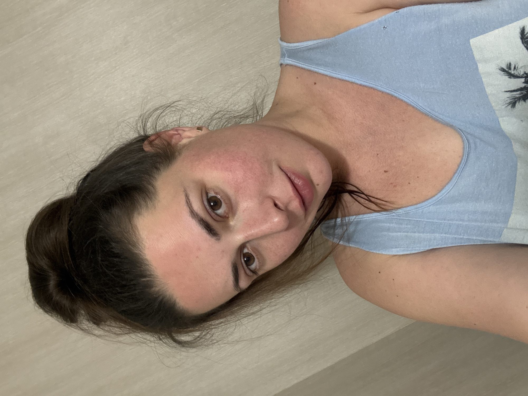 A woman with brown hair pulled into a bun is seen in a close-up, head-and-shoulders shot. She is wearing a light blue tank top. Her skin appears flushed on her cheeks and forehead. The background is a textured, neutral-colored wall. The image is taken from a slightly upward angle.