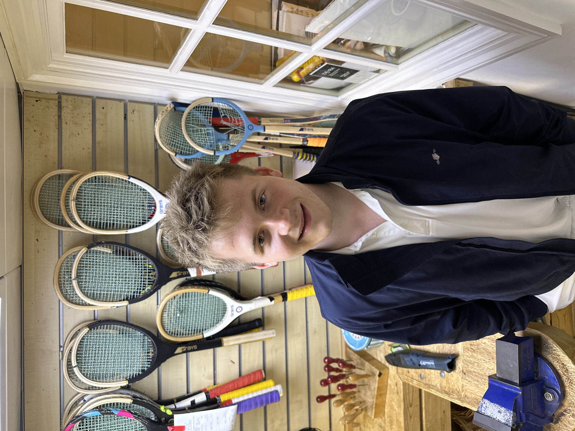 A medium shot features a blond-haired man in a navy jacket and white collared shirt, positioned in front of a wall adorned with vintage sporting equipment. The man is smiling slightly and looking toward the camera.
Behind him, a collection of old tennis rackets and badminton rackets are mounted on a paneled wall. A window is visible in the upper part of the frame.
To the right of the man, a workbench includes a blue vice, hand tools, and other miscellaneous items. The scene suggests a workshop or storage area related to sports equipment, possibly in London, United Kingdom.
