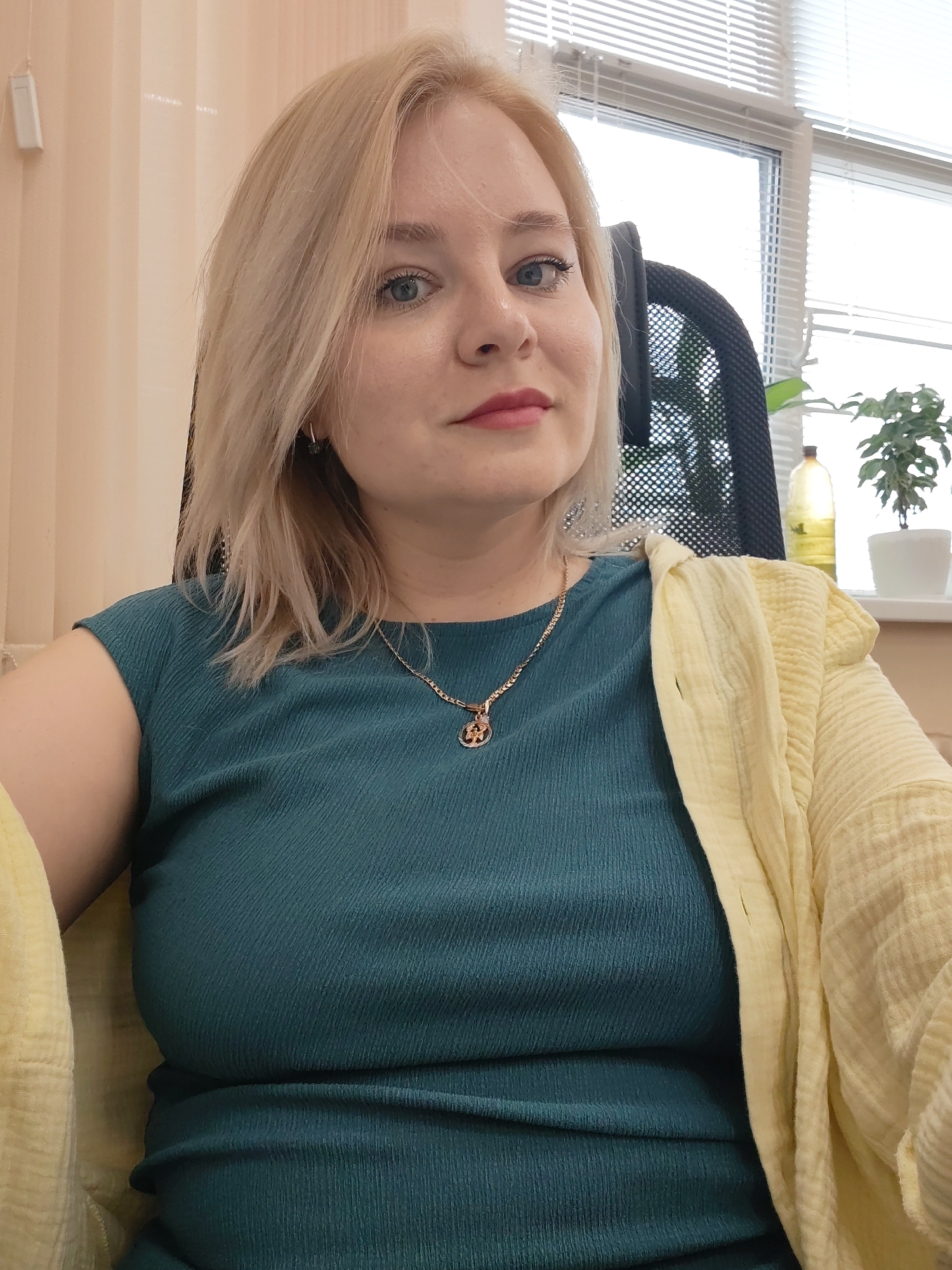 A blonde woman sits in a black mesh office chair, wearing a teal dress and a pale yellow cardigan draped over her shoulders. She is facing the camera. She has a gold necklace with a pendant. The background shows a window with blinds, a houseplant in a white pot, and a bottle of oil. The location is identified as Izhevsk, Russia.