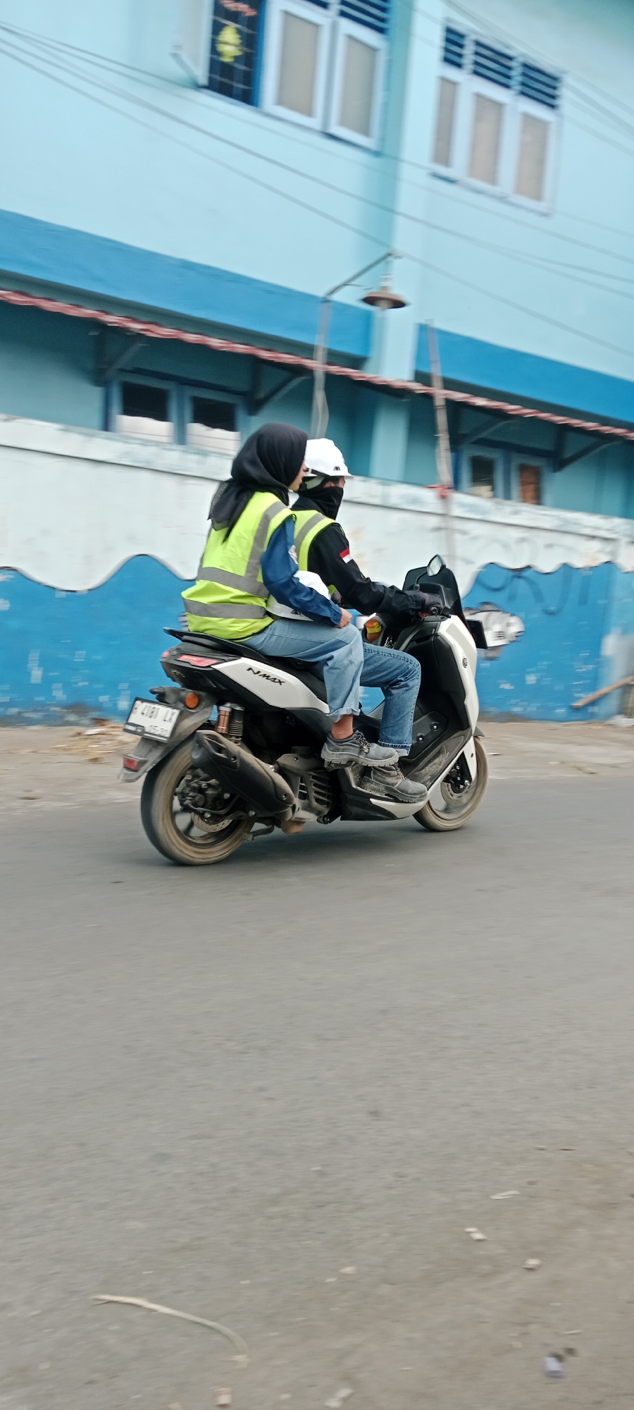 Two individuals are riding on a white scooter in Purwomartani, Indonesia. The person in the back, wearing a black hijab and a neon yellow vest over a blue shirt, is seated behind the driver. The driver, wearing a white helmet and a dark jacket with an Indonesian flag patch, is operating the scooter. Both individuals are wearing blue jeans. The scooter has a license plate with the number "R 4181 LX". The background features a light blue building with white-framed windows and a blue decorative wave pattern along the lower facade. A street lamp is visible mounted on the building. The scooter is on an asphalt road.