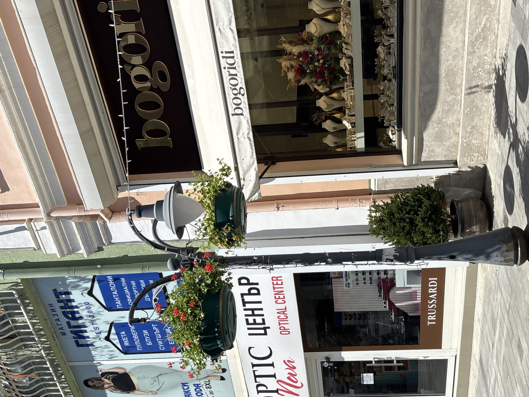The image shows a street scene in the city of Burgas, Bulgaria, specifically on Bogoridi Street. In the foreground, a lamppost with two hanging baskets of red flowers is prominently featured. To the left, a storefront for "OPTIC CENTER Nova" is visible, with the text "OPTICAL CENTER" below it. Further to the left, a sign for a clothing store with the Bulgarian word "ГИГАНТ" (GIGANT) is seen, along with images of clothing items. To the right of the lamppost, a jewelry store with the name "bogoridi" displayed on its awning and facade is evident. The window display of the jewelry store showcases various necklaces, earrings, and other jewelry pieces, along with decorative floral arrangements. The storefront for "TRUSSARDI" is also visible on the lower part of the image, to the left of the jewelry store. The scene is captured on a sunny day, with shadows cast on the pavement.