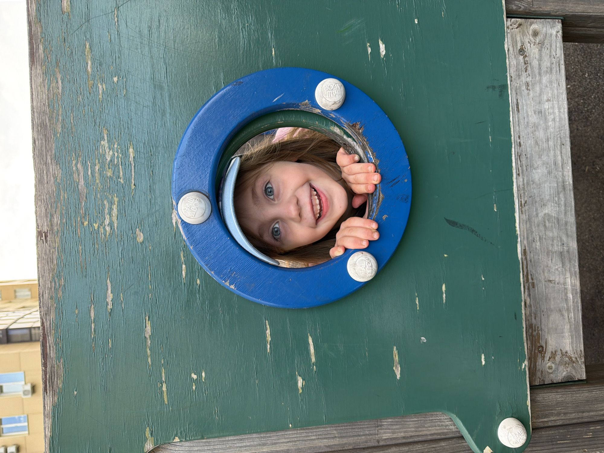 The image shows a young girl peeking through a circular opening in a weathered green wooden structure. The girl, wearing a light blue baseball cap, has wide blue eyes and a big smile. Her hands are visible, resting on the edge of the opening. The wooden structure appears to be part of a playground, with peeling green paint and some visible wood grain. In the background, the upper floors of a building with yellow walls and several windows are visible, suggesting an urban setting. The overall scene appears to be outdoors on a cloudy day. The image does not provide specific location information within the city of Perm, Russia.