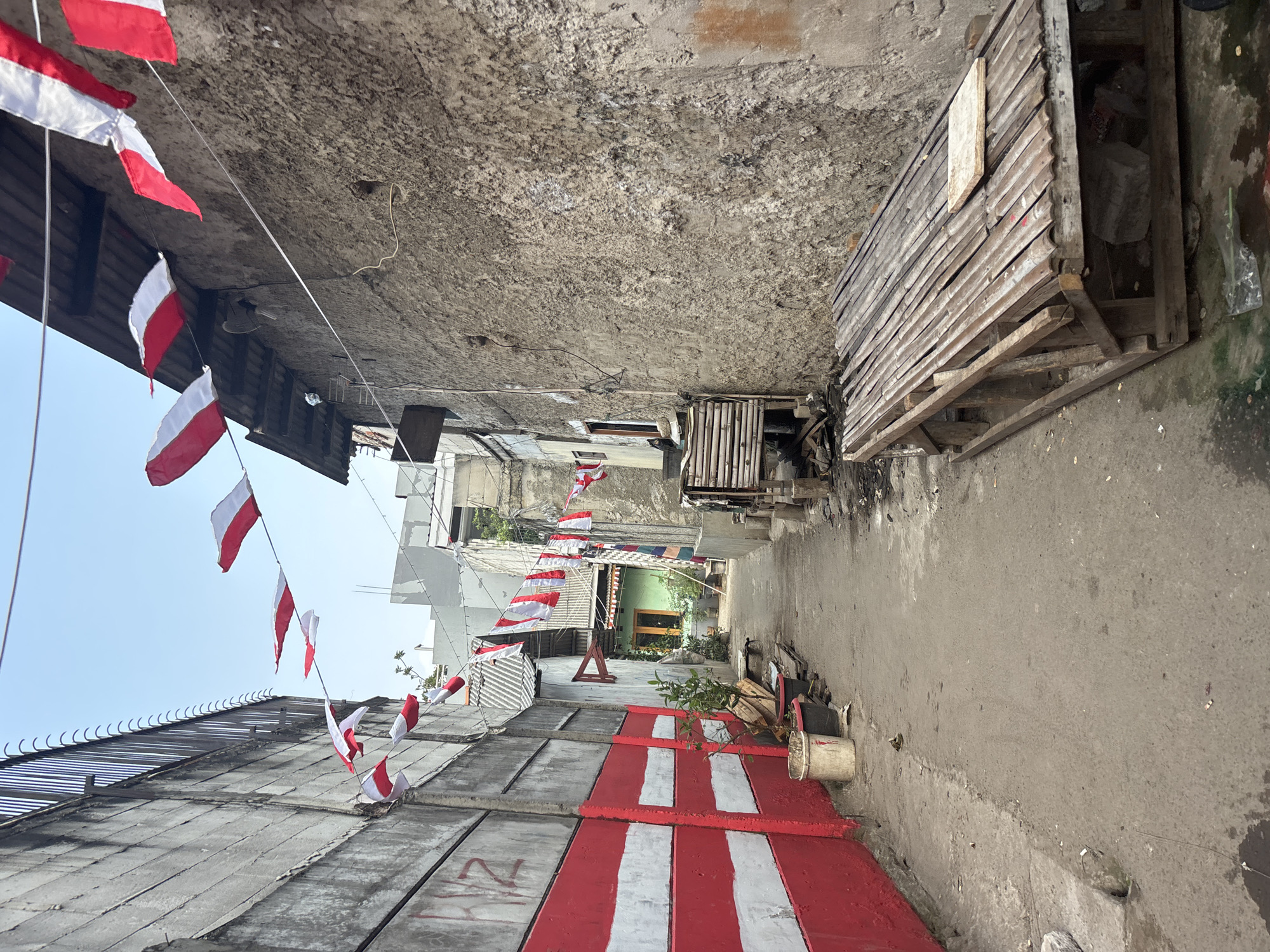 The image captures a narrow alleyway in Bekasi, Indonesia, decorated with a string of red and white flags, suggesting a celebration or national event. The alley is flanked by textured, aged concrete walls, one of which is boldly painted with horizontal red and white stripes. On the left side of the alley, a tall, grey concrete wall with a barbed wire fence atop it runs alongside. Various items are scattered throughout the alley, including buckets, potted plants, and pieces of wood. In the foreground, a rustic wooden structure with a corrugated metal roof lies on the ground, and more wooden planks are visible nearby. The overall scene portrays a common urban environment in Bekasi, with evidence of everyday life and festive decorations.