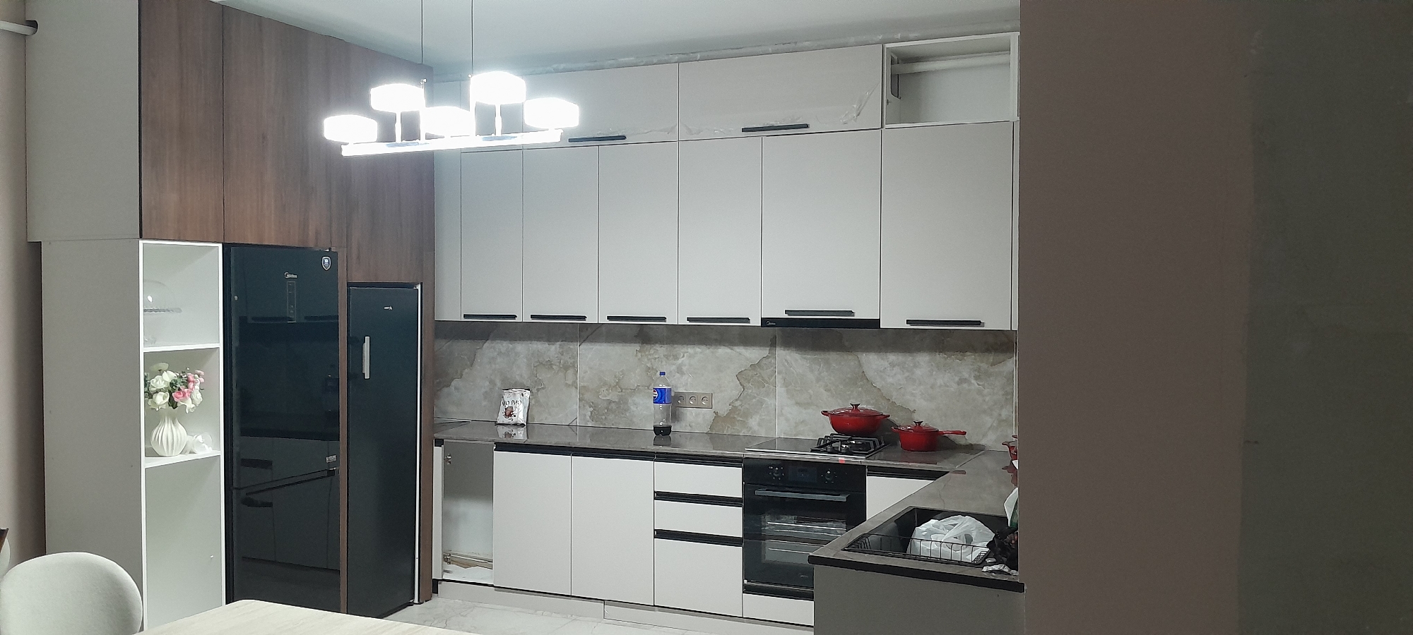 The image depicts a modern kitchen interior. The kitchen features white cabinets with black handles, a dark countertop, and a marble-patterned backsplash. A black refrigerator is visible on the left, next to a white shelving unit containing a vase with flowers. Above the counter, there are several upper cabinets and a contemporary light fixture with multiple bulbs. A stove with an oven is built into the counter, with two red pots on top. There is also a bottle of water and a package of food on the counter. The overall aesthetic is clean and minimalist. The location is identified as a kitchen in Kyzyl-Asker, Kyrgyzstan.