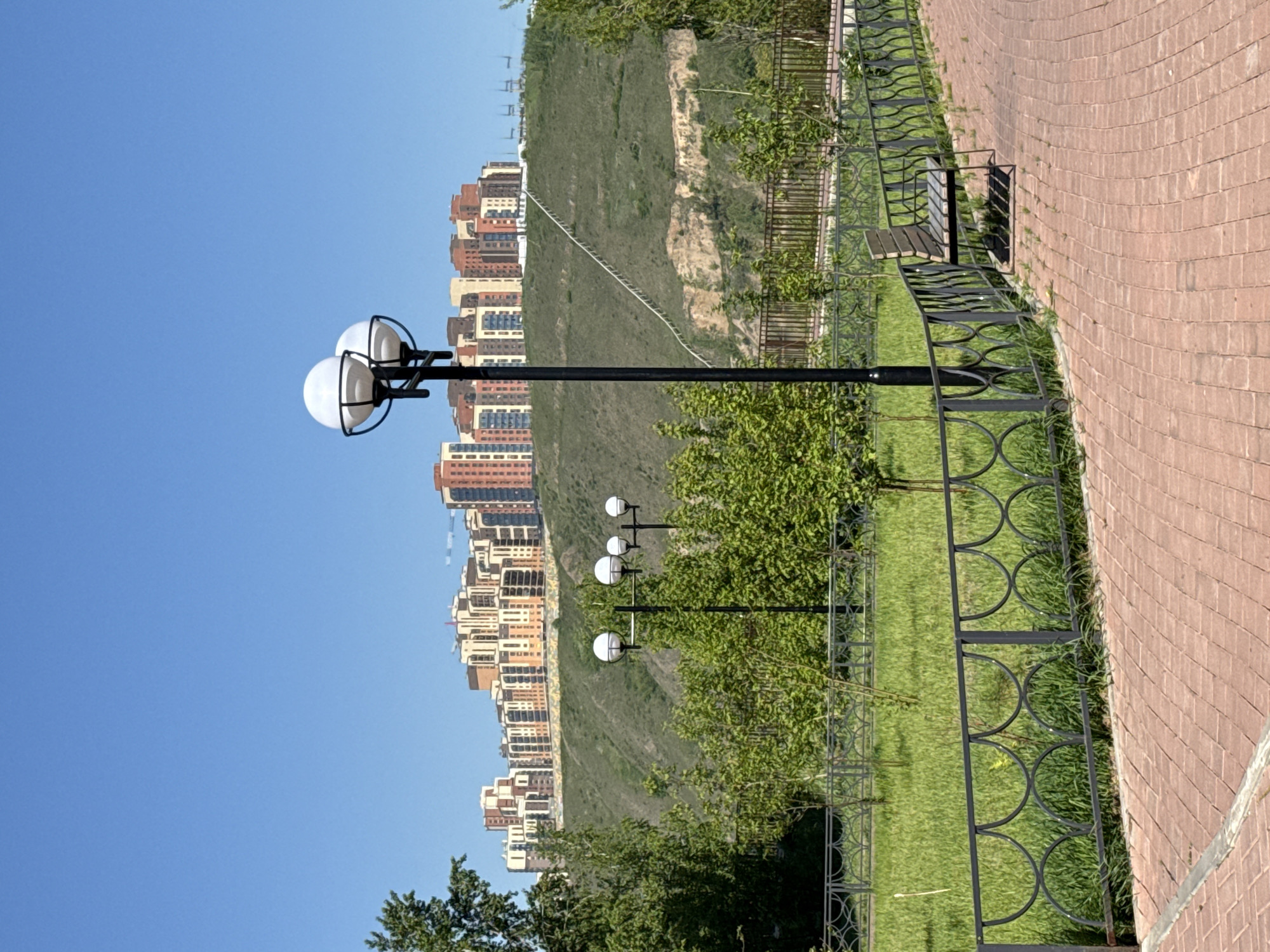 This image captures a bright, sunny outdoor scene in Krasnoyarsk, Russia, featuring a paved walkway, a park area, and distant residential buildings set against a hill.
In the foreground, a reddish-brown brick-paved path curves gently from the bottom right towards the top left. The bricks are laid in an arching, tessellated pattern. To the left of the path, a decorative black metal fence with repeating circular and semi-circular motifs borders a neatly manicured green lawn. A wooden park bench with a black frame is visible next to the fence on the right side of the image, casting a clear shadow.
Several black lampposts with white spherical lights stand on the grassy area. One prominent lamppost with two lights is centered in the mid-ground, while others with two and three lights are further back. Beyond the fence and grass, a large, verdant hill rises, covered with green vegetation, but also showing patches of exposed earth. A thin, light-colored line, possibly a pipe or cable, ascends diagonally across the hillside.
Atop and behind the hill, a skyline of modern, multi-story residential buildings is visible. These buildings feature varied facades in shades of brown, red, and beige, with numerous windows. The sky above is a clear, vibrant blue, indicating excellent weather conditions. The strong sunlight casts crisp shadows, suggesting it is either late morning or early afternoon. No people or visible text are present in the image. The scene conveys a sense of urban tranquility, offering a pleasant recreational space with a view of the city and natural landscape.