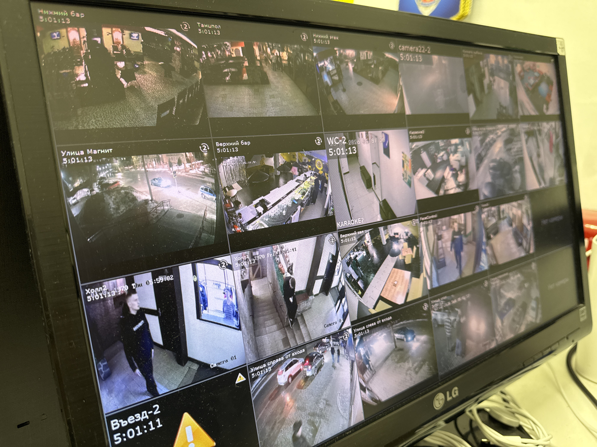 A black LG monitor displays a 4x6 grid of surveillance camera feeds, collectively showing various locations within a complex and its immediate surroundings in Chaikovsky, Russia. Most feeds are timestamped "5:01:13," with some showing an incorrect system date like "1970 Thu 01:59:02."
The feeds include:
* **Interior views:**
* Multiple bar areas ("Нижний Бар," "Верхний бар") showing counters with bottles, tables, chairs, and several individuals standing or seated.
* A dance floor ("Танцпол") with individuals present.
* Hallways and common areas ("Холл," "Нижний этаж," "Верхний зал") where individuals are seen walking, including one person in dark clothing with a white word on their chest.
* A restroom ("WC-2") showing stalls.
* A karaoke room ("Karaoke2") with seating.
* **Exterior views:**
* Street scenes ("Улица Магнит," "Улица справа от входа," "Улица слева от входа") at night, depicting parked and moving vehicles, streetlights, buildings, and several pedestrians on sidewalks. The ground shows evidence of snow or slush.
* One feed labeled "Въезд-2" (Entrance-2) partially displays a yellow triangular warning sign with an exclamation mark.
The overall environment suggests a commercial establishment like a bar, club, or restaurant, with both indoor activity and exterior street monitoring.