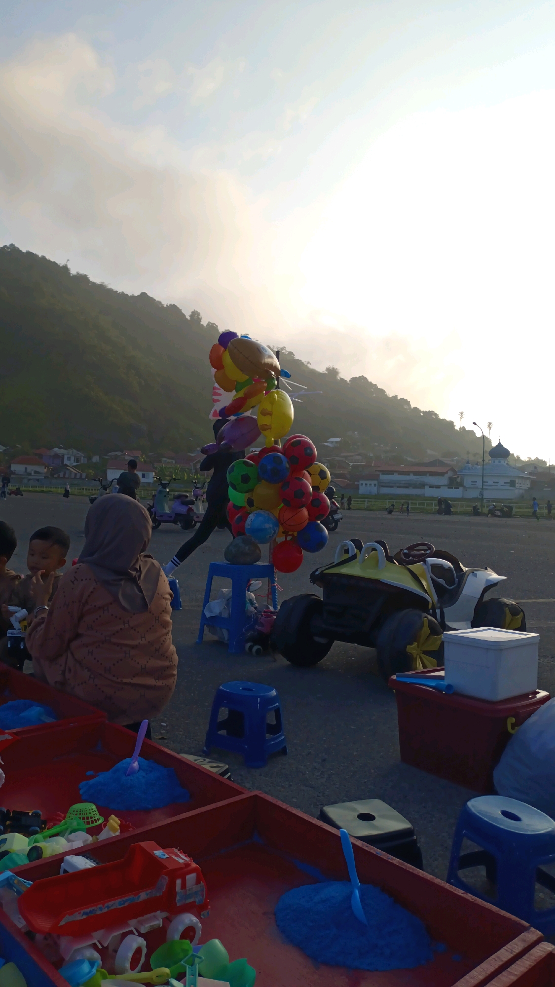The image depicts a street scene in Padang Panjang, Indonesia, likely near a coastal area or a recreational park given the presence of a sandy play area and a large body of water in the distance. In the foreground, a red sandbox is filled with blue sand and children's toys, including a red dump truck. To the right of the sandbox, a yellow and black ride-on toy car is parked.
In the middle ground, a person dressed in dark clothing stands next to a display of colorful balloons, many of which are shaped like soccer balls and footballs. The balloons are attached to a stand and are being held up by the person. Several blue plastic stools are scattered around the area.
Further back, a woman wearing a hijab and a patterned brown top is seated, facing away from the camera, with two young children beside her. In the background, a hazy mountain range is visible, with a town nestled at its base. Buildings, including a white structure with a blue dome, are discernible. Various individuals and motorcycles are also present in the background, suggesting a public space. The sky is bright and overcast, indicating it might be early morning or late afternoon.